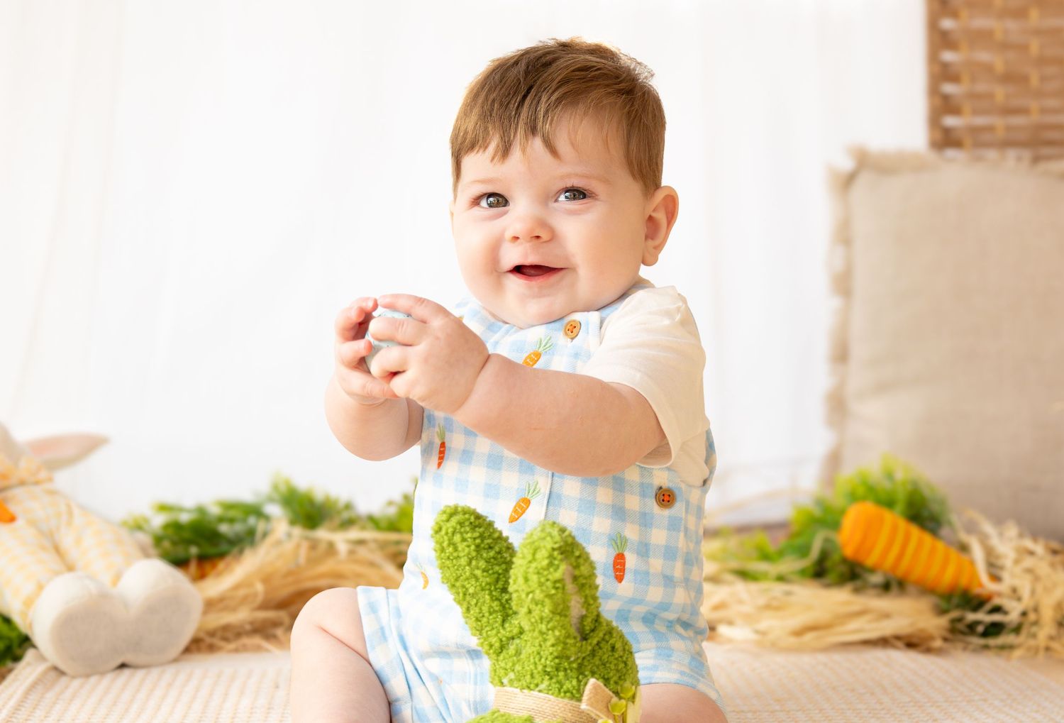 Smiling baby in blue gingham romper with bunny applique sitting among Easter decorations and carrots.