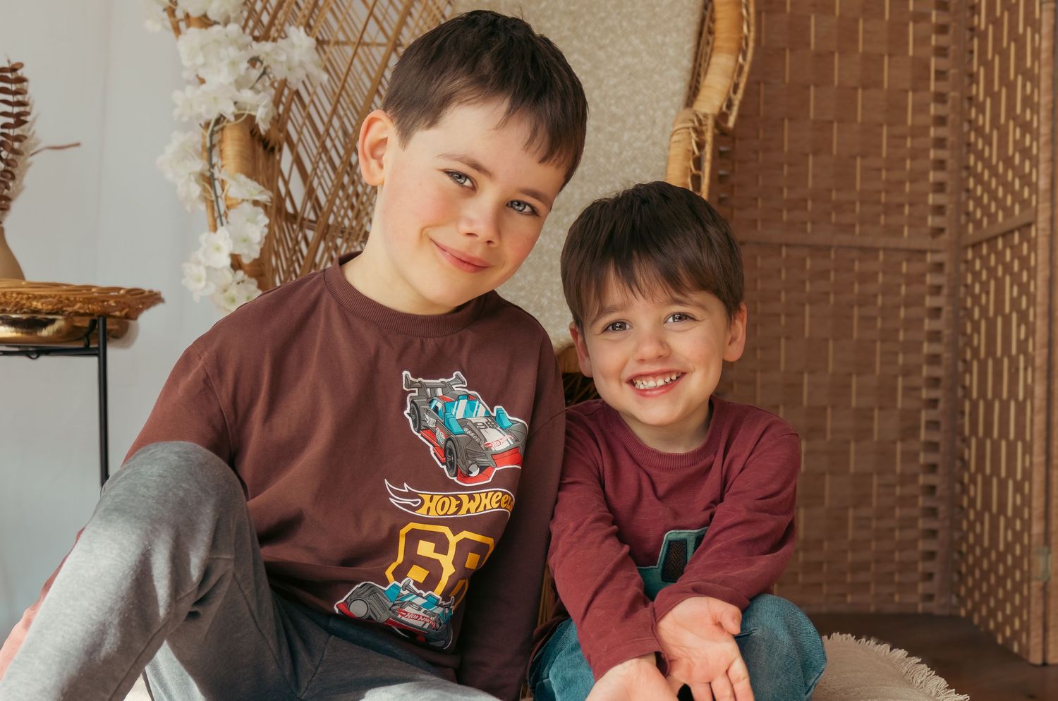 Two young boys smiling and sitting together in a cozy wicker chair, wearing matching burgundy shirts.