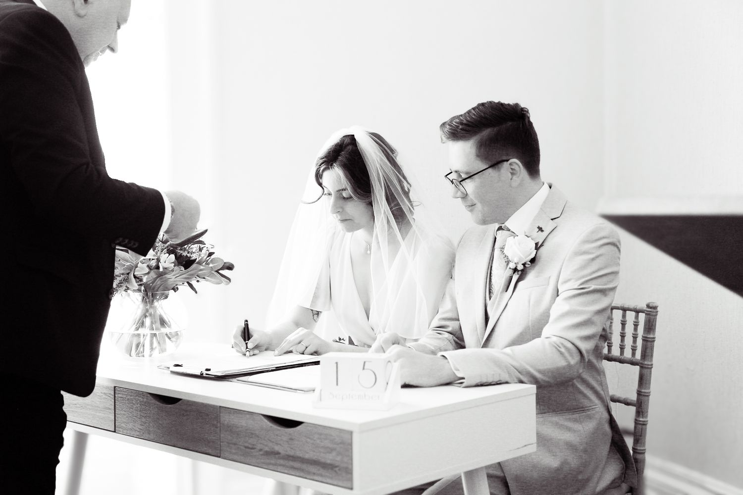 Black and white photo of a couple signing marriage documents at a white table.