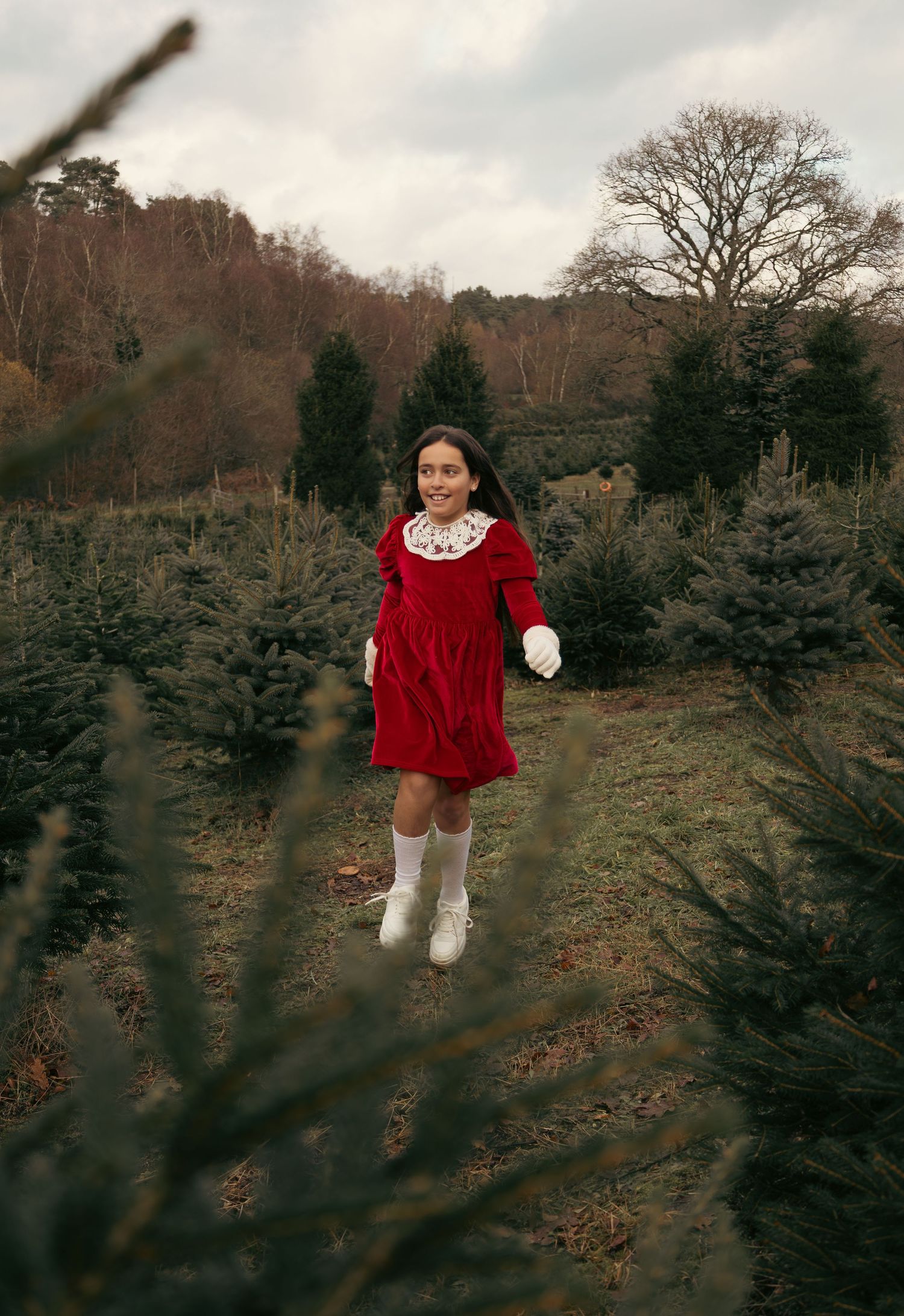 Someone in a red velvet dress with white trim stands in a field surrounded by pine trees.