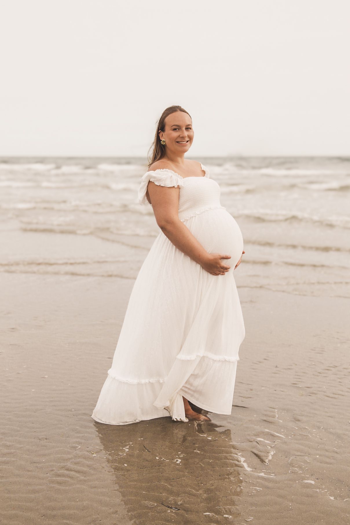Expectant mother twirls playfully in white dress at water's edge.