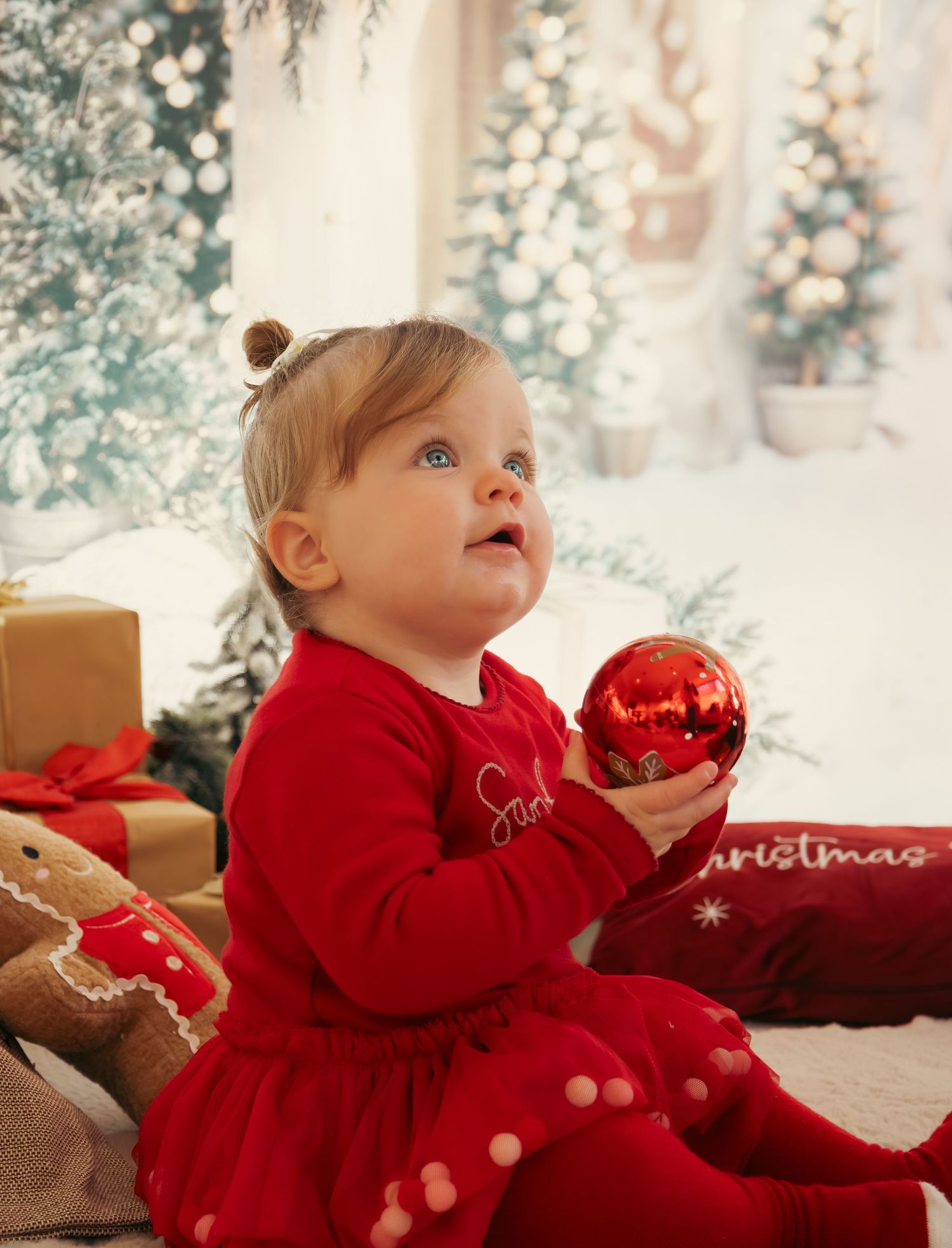 Baby in red holiday outfit holding shiny red ornament ball surrounded by Christmas decorations.