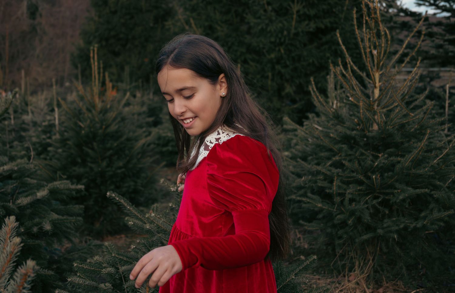 Person in red velvet coat stands among Christmas trees in winter setting.