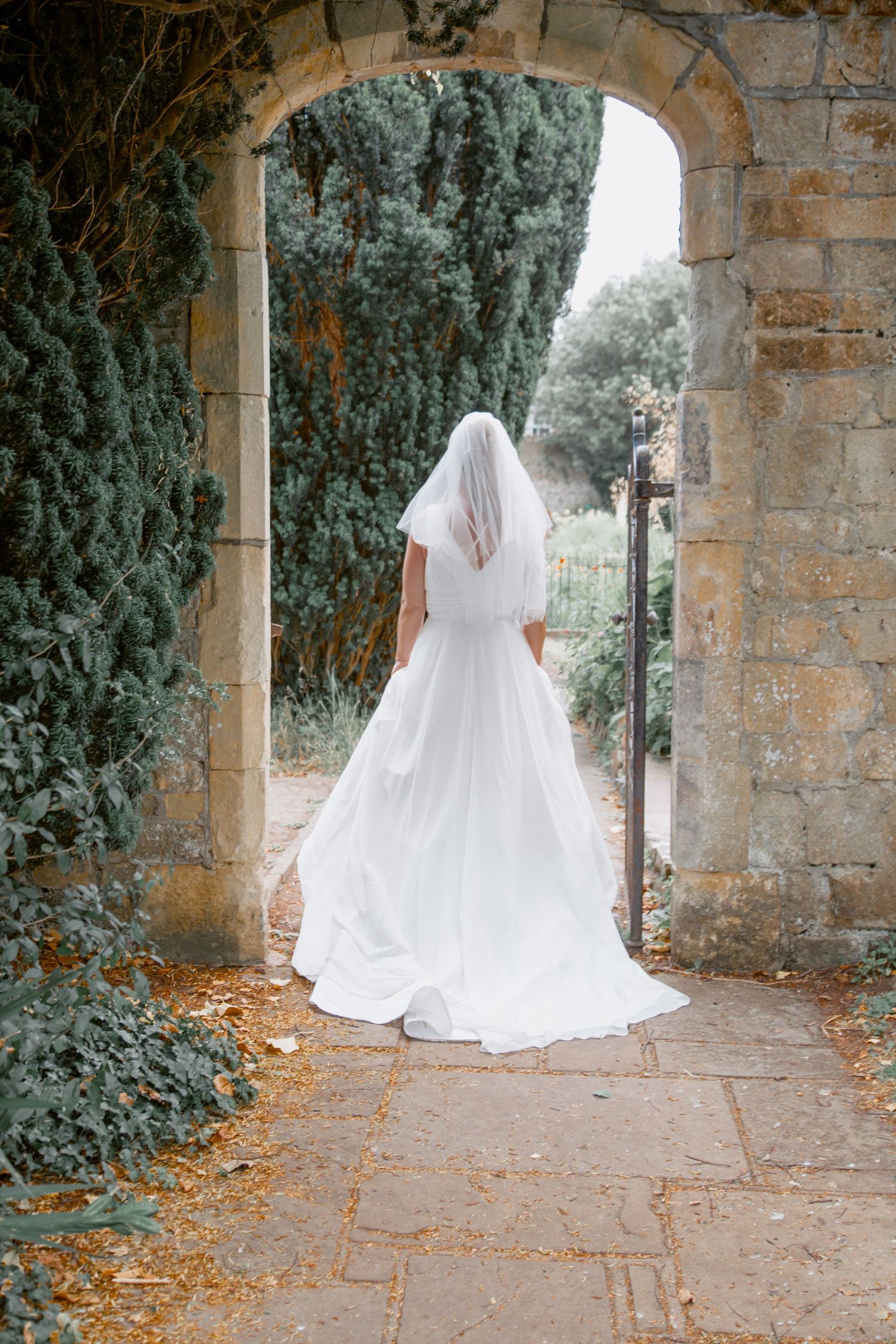 A bride in a flowing white wedding dress poses in a stone archway surrounded by greenery.