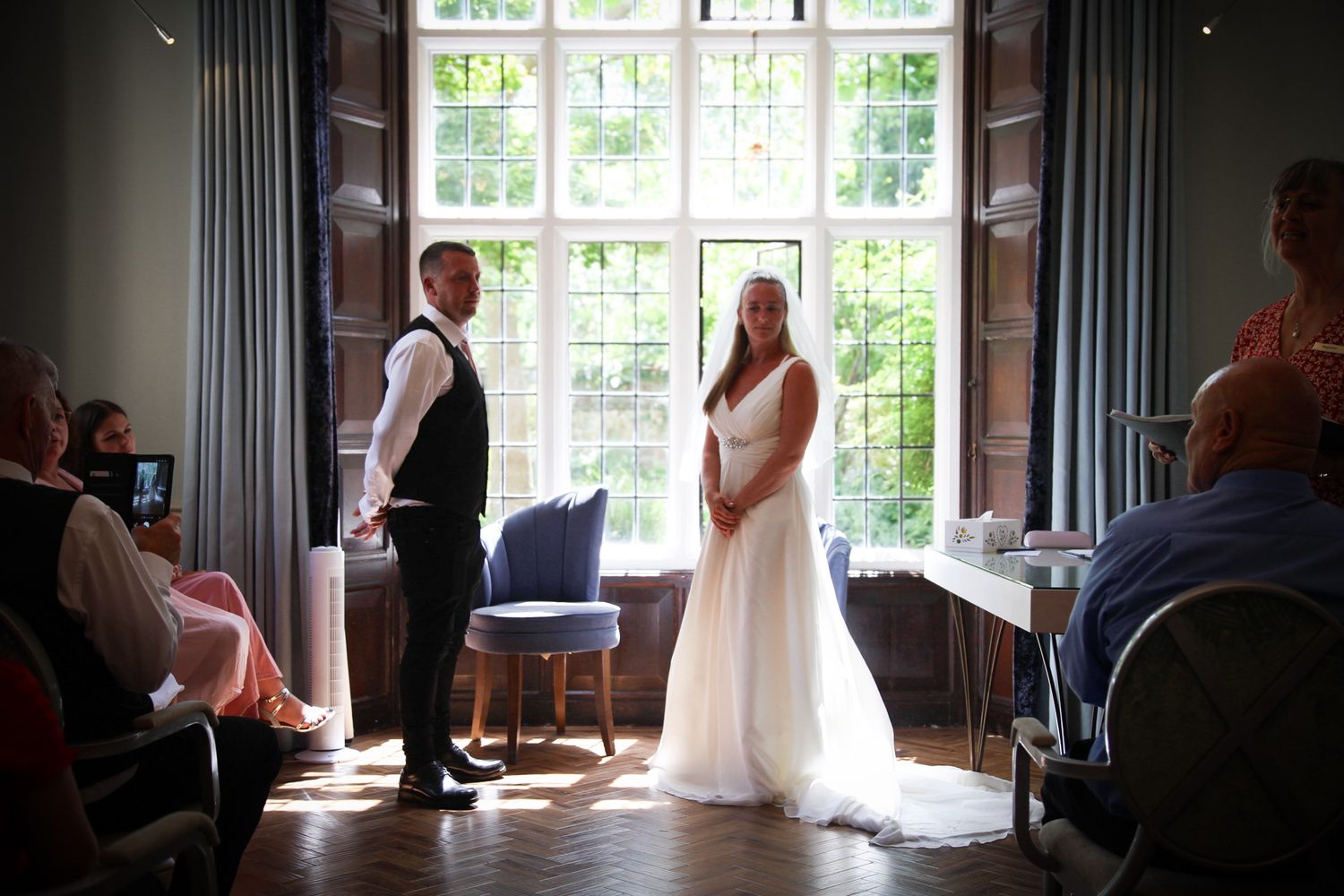 A wedding ceremony takes place in front of large windows with natural light streaming in.