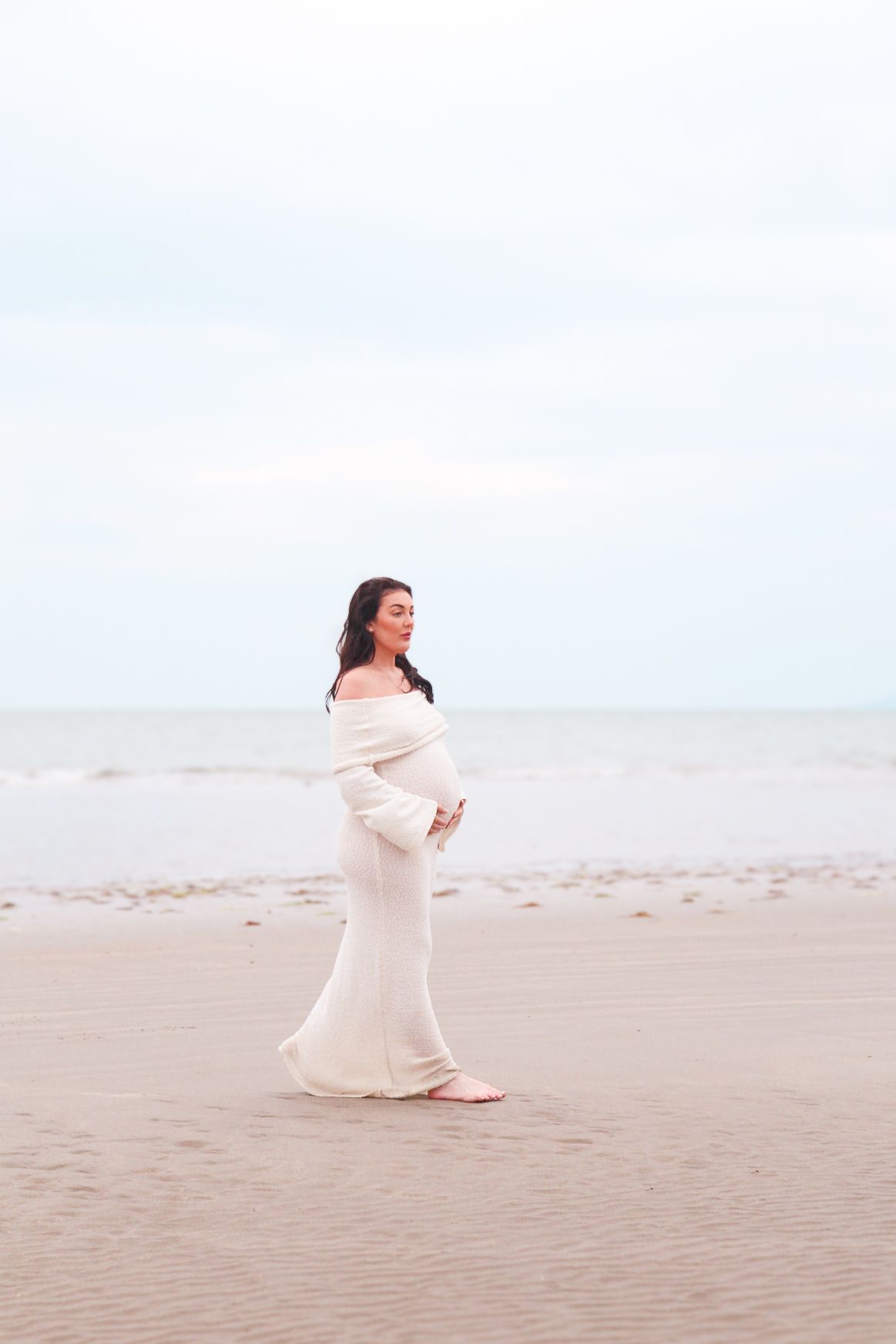 A graceful beachside maternity photo featuring a cream-colored dress against a misty coastal backdrop.