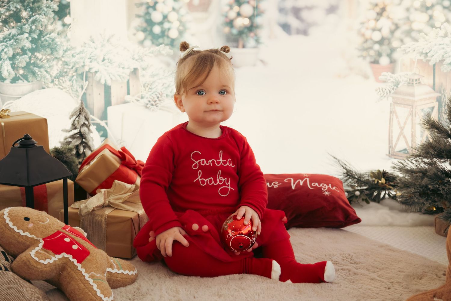Small child in red Christmas outfit sitting among wrapped presents and holiday decor.