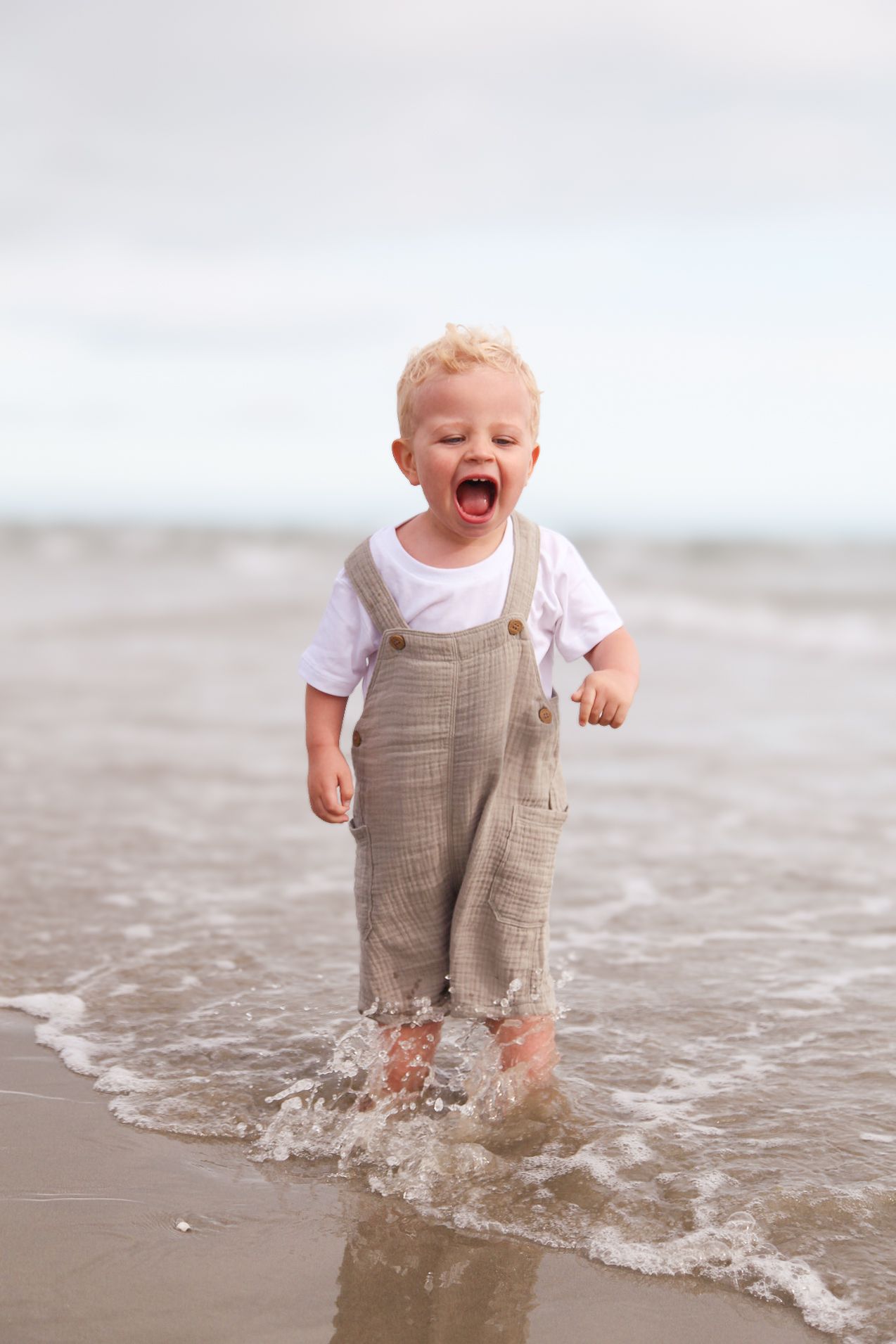 Happy toddler splashing in shallow ocean waves wearing tan overalls.