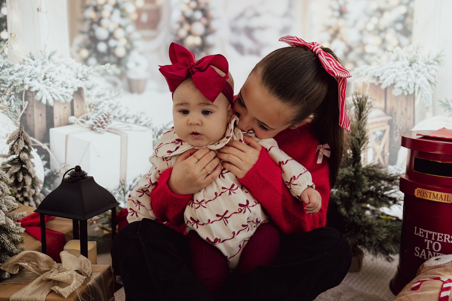 Mother in red sweater holds baby in reindeer onesie with red bow, posing in festive Christmas holiday photo setting.