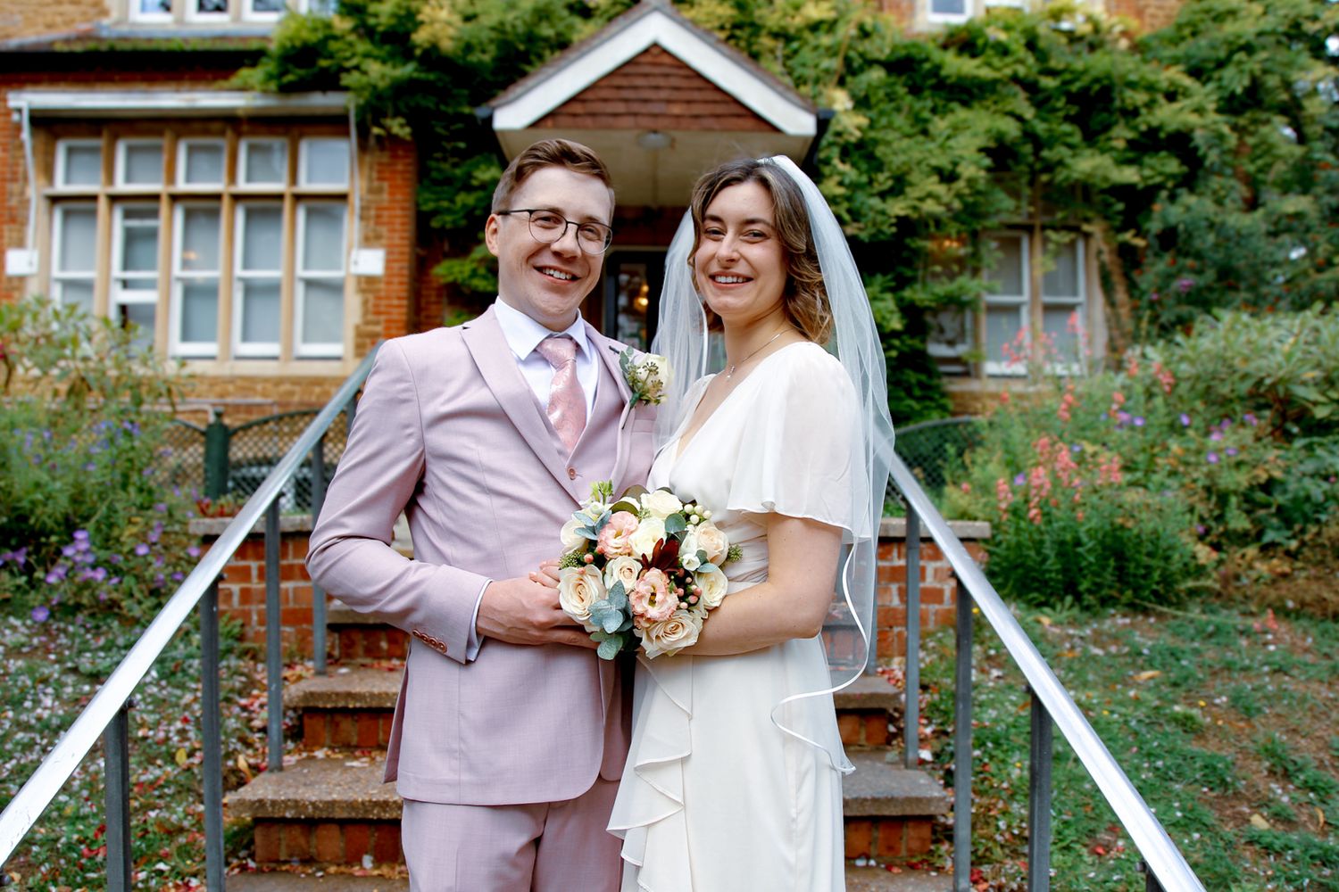 A couple in a pink suit and white dress share a romantic moment in front of a brick building with greenery.