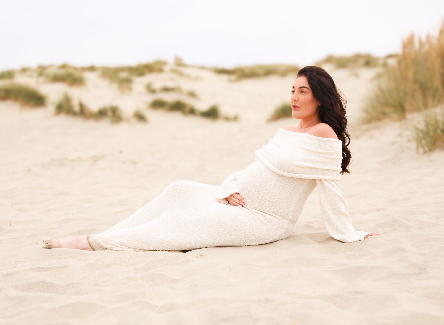 Expectant mother in flowing white dress reclines peacefully on sandy beach.
