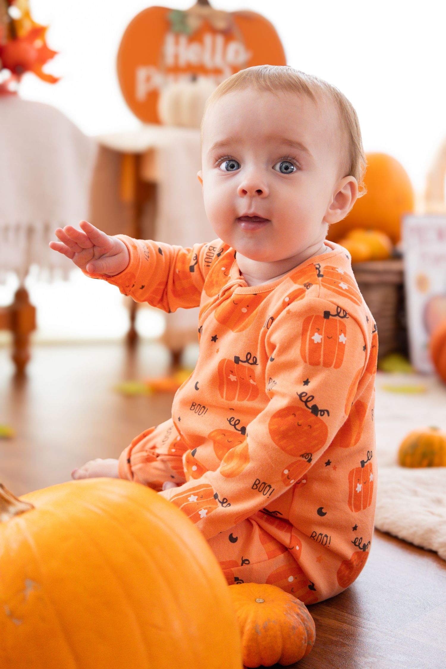 Baby in orange pumpkin-print outfit sitting next to large pumpkin with fall decorations.