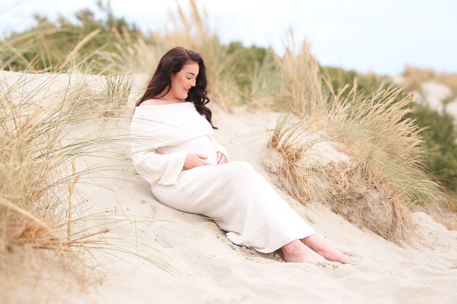 Woman in white dress poses gracefully among coastal sand dunes and sea grass.