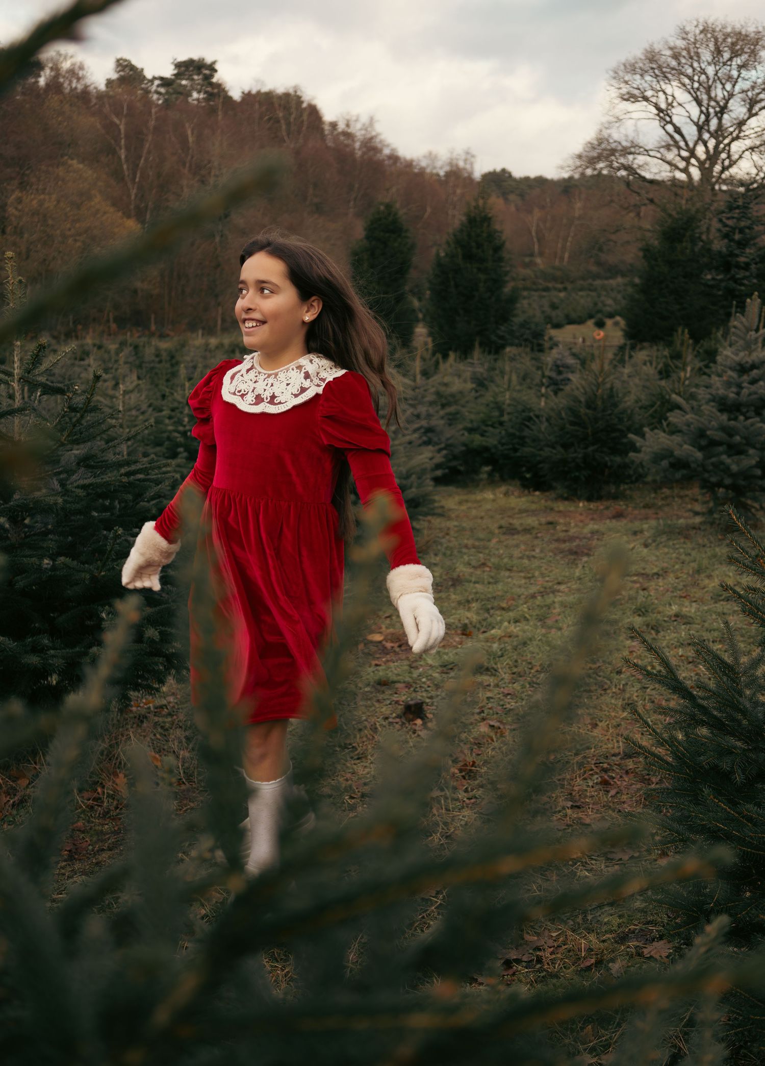 Someone wearing a red dress with white collar twirls playfully among evergreen trees.