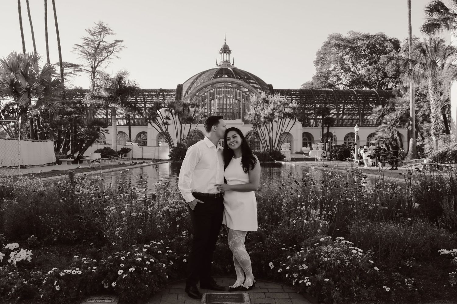Black and white portrait of a couple standing before a majestic conservatory with blooming flowers.