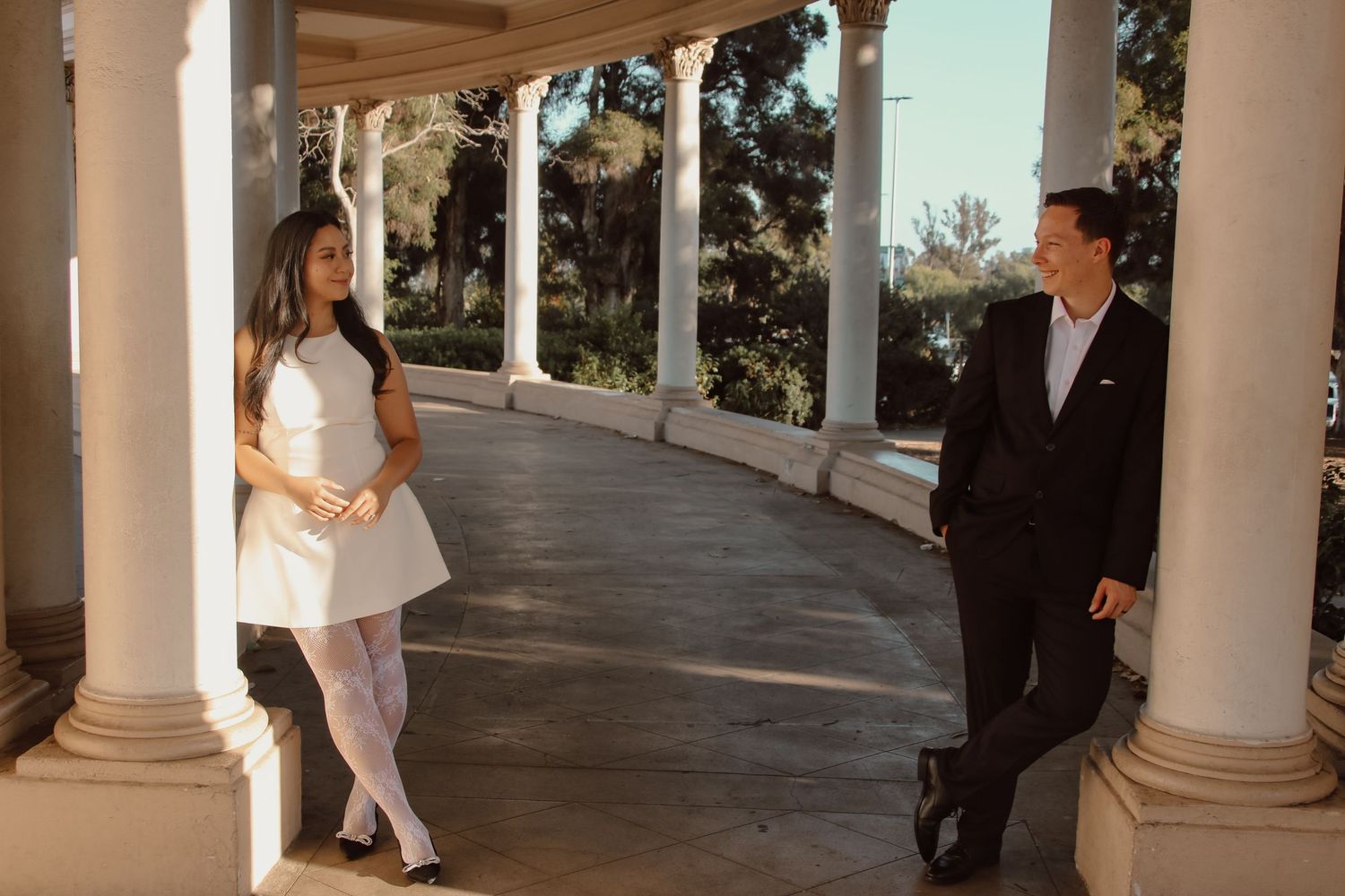 A couple in contrasting formal wear poses along a curved colonnade walkway.