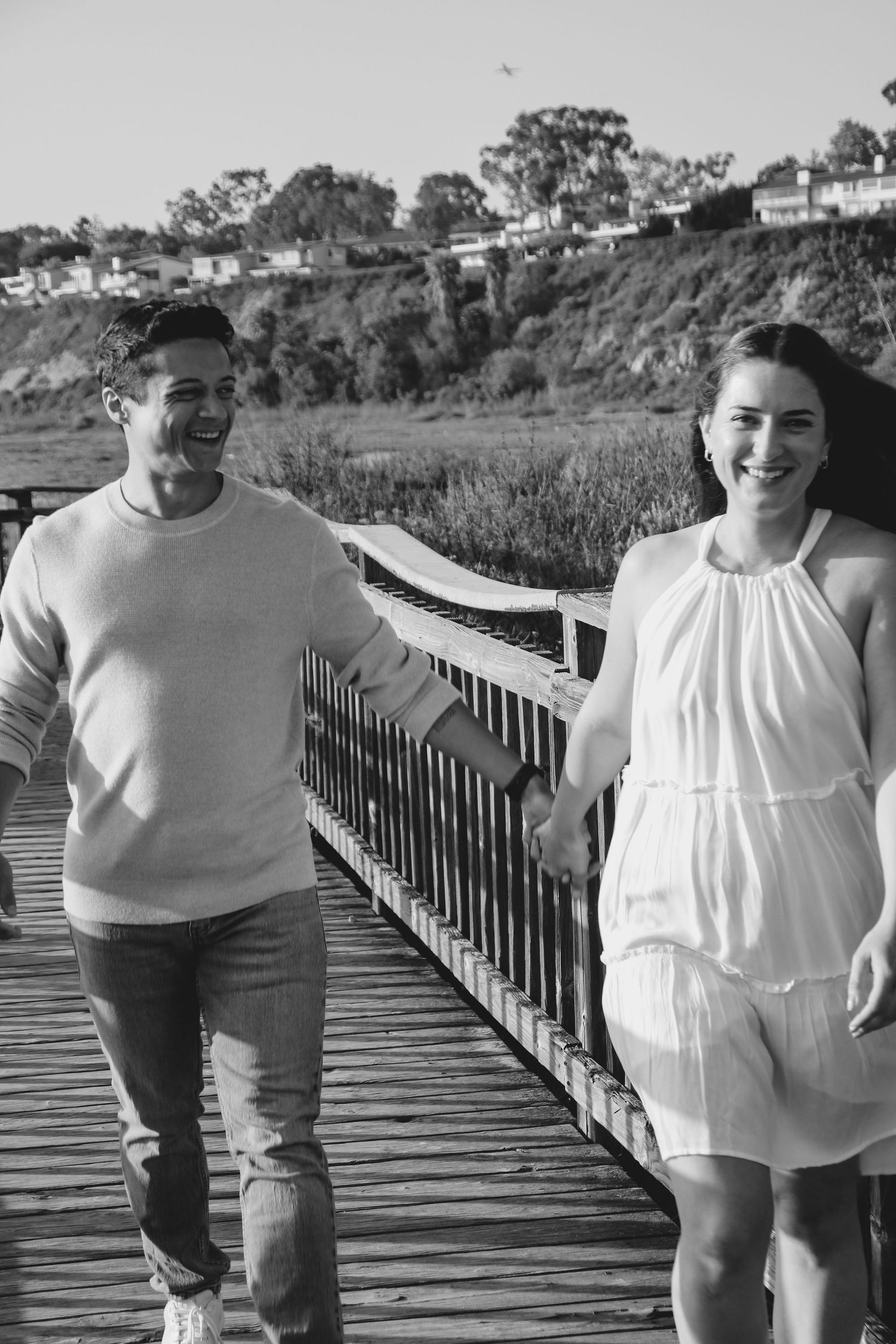 Black and white photo of two people walking and laughing together on a boardwalk near the coast.