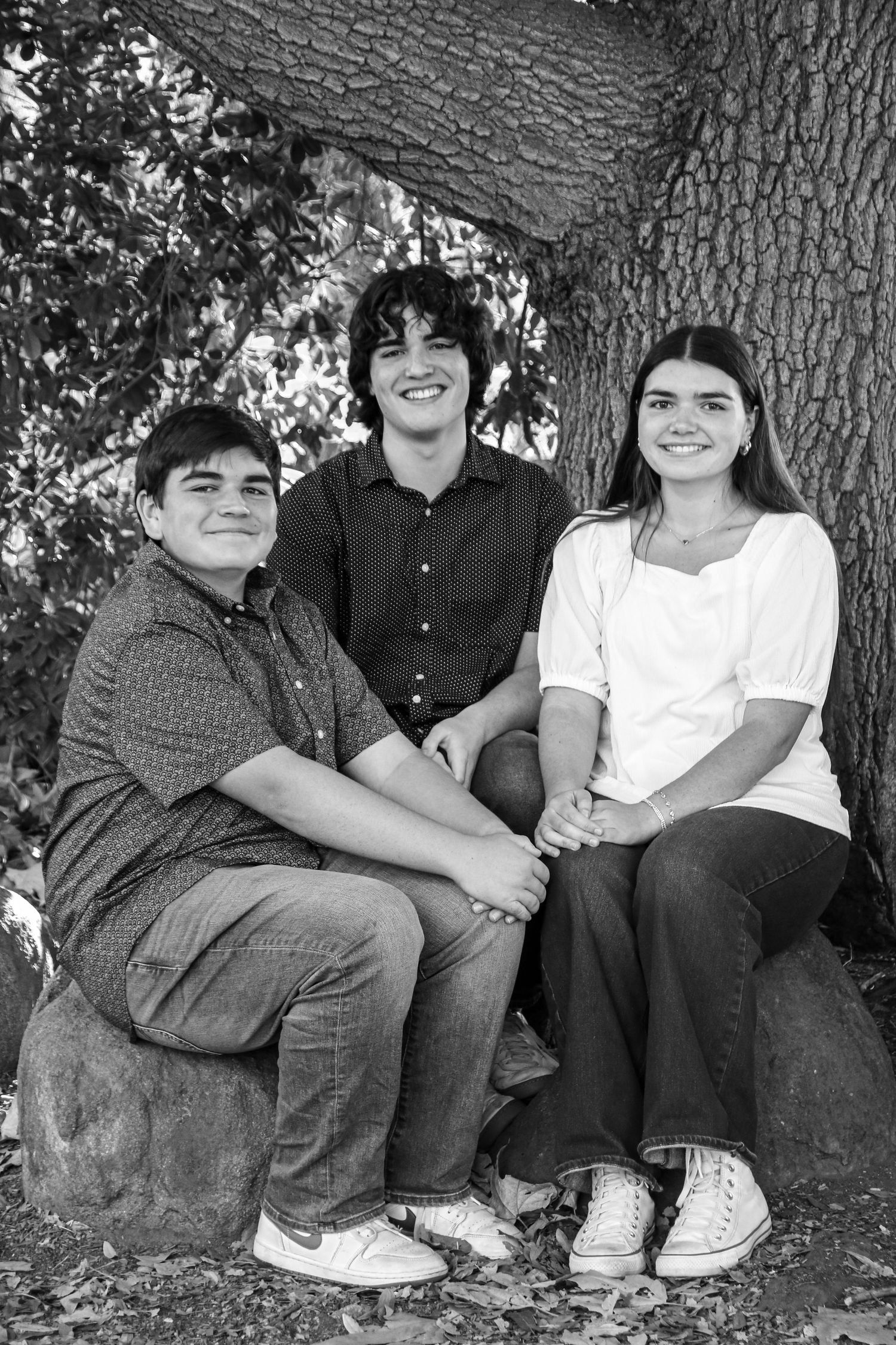 Three young people sitting on rock by tree trunk in casual black and white portrait.