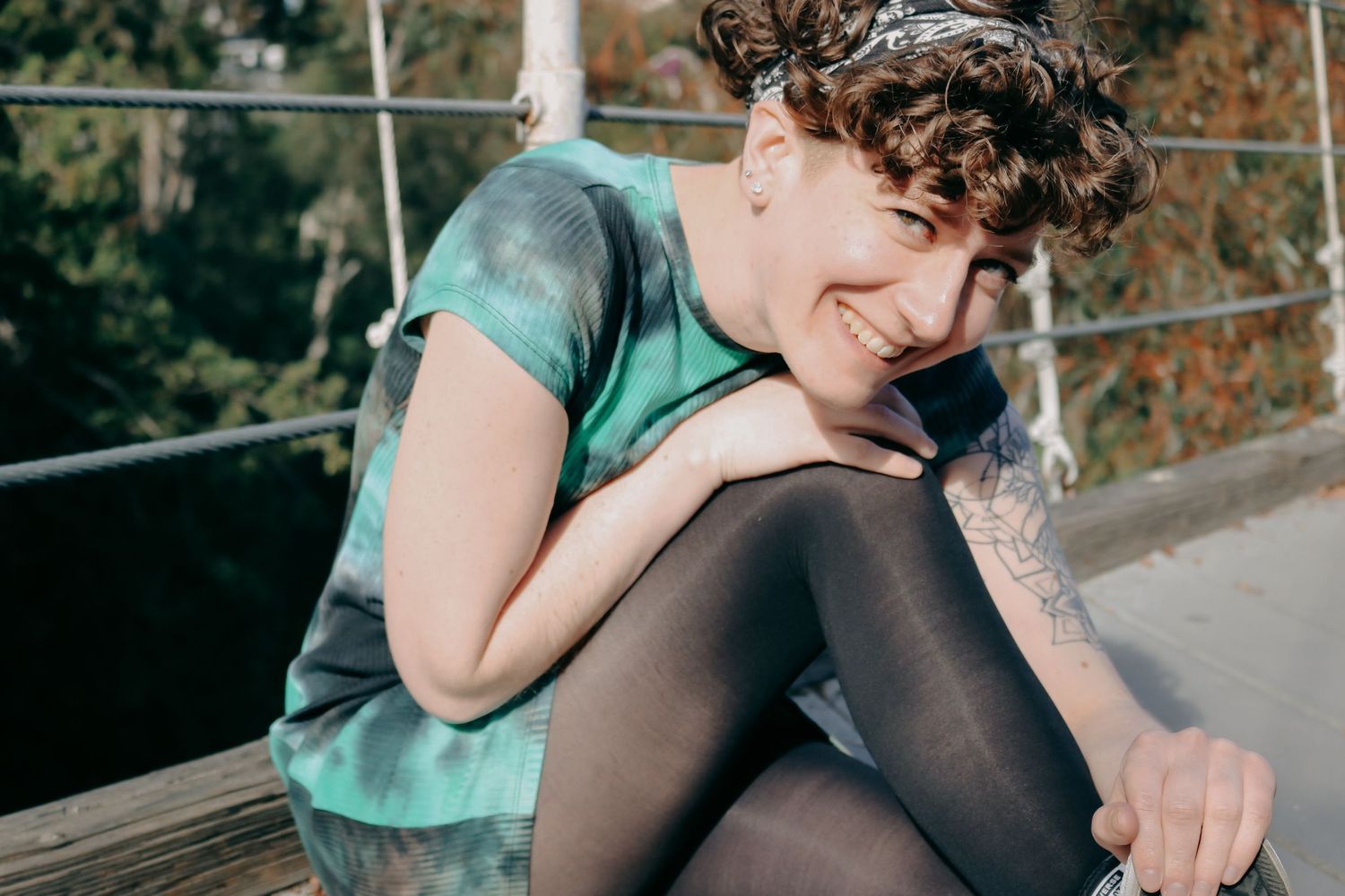 Person in black dress sits on rock in nature setting with curly hair and tattoo sleeve.