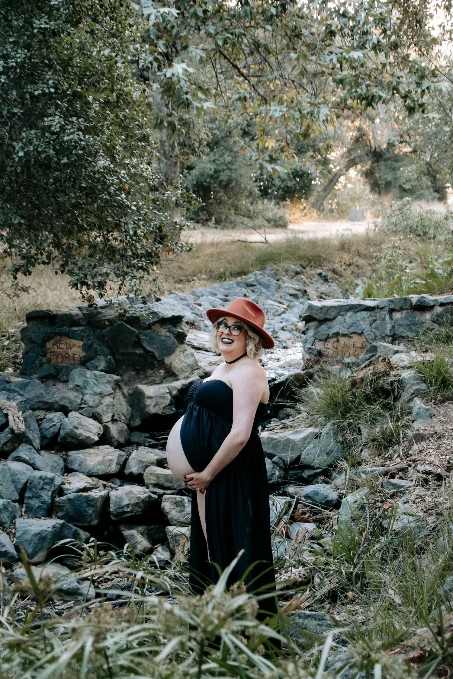 A scenic maternity photo with stone wall backdrop showing a figure in black dress and rust-colored hat.