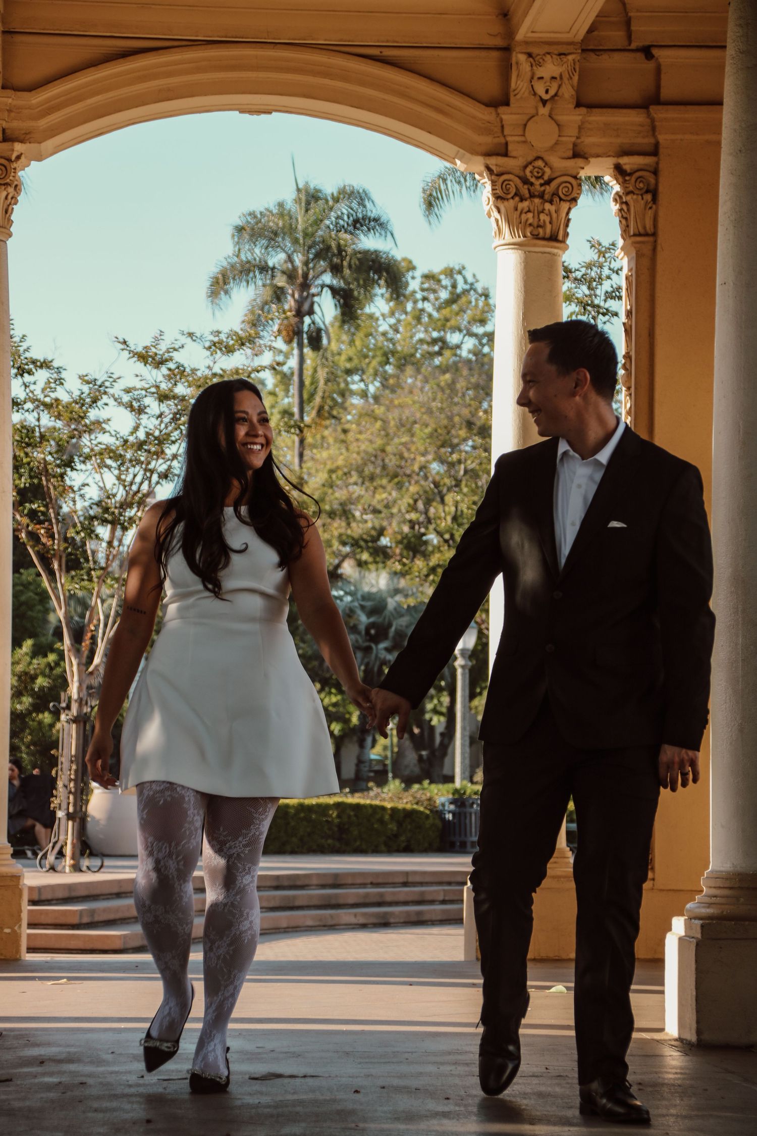 A stylishly dressed couple shares a joyful moment while walking through an elegant portico.