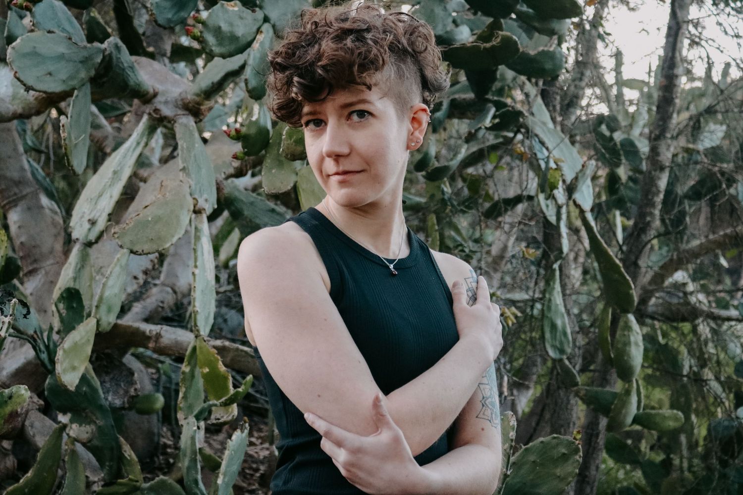 Natural light portrait against desert plants with arms crossed wearing black tank top.