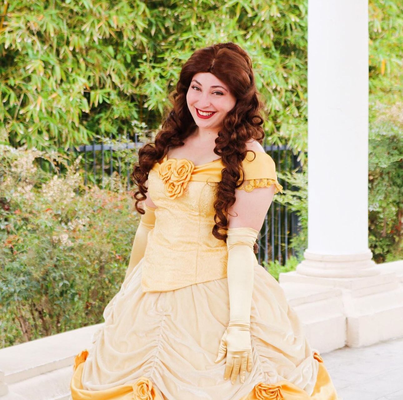 A cosplayer in an elegant yellow ballgown poses gracefully by a white column.