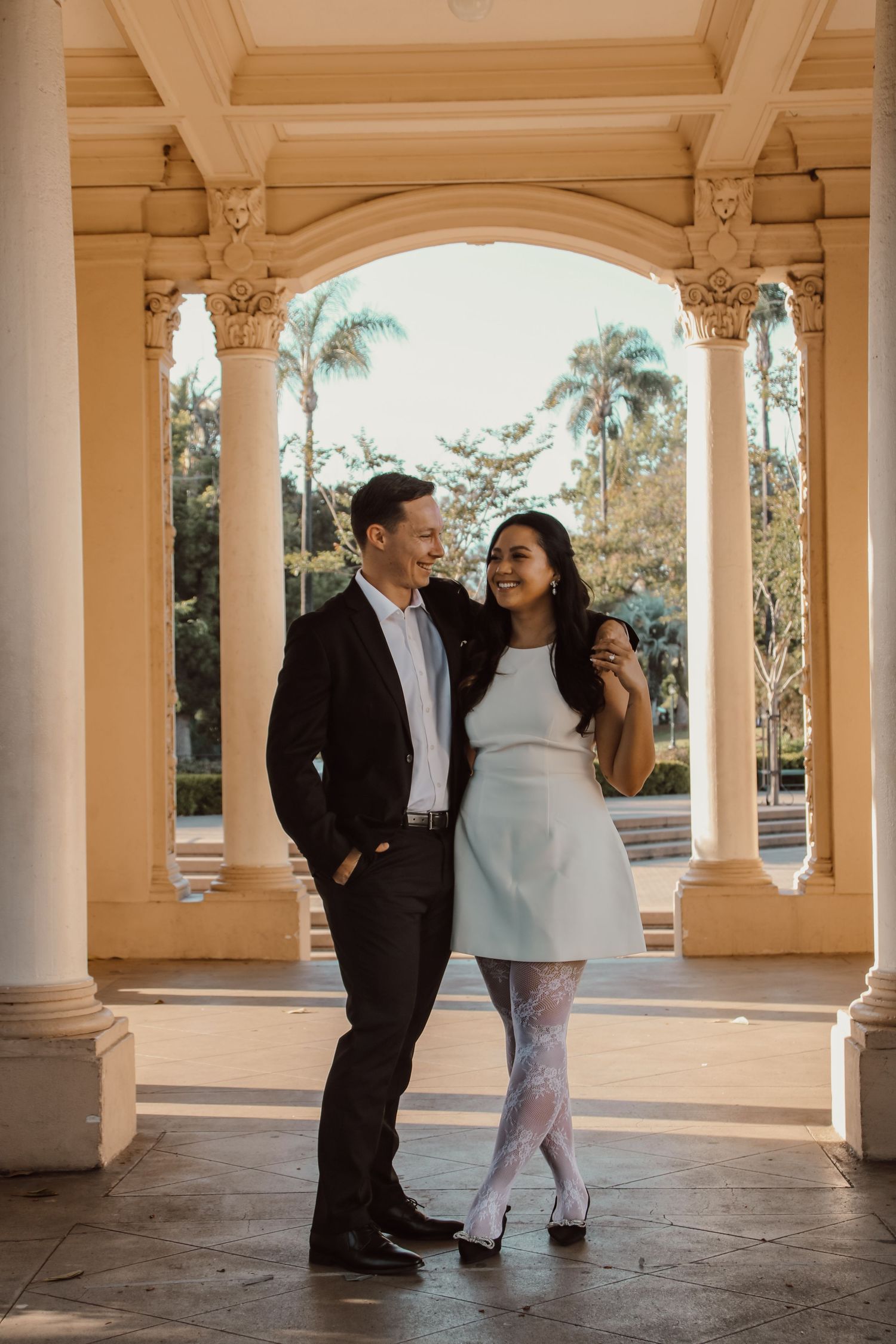 Two people share intimate moments in a sunlit classical colonnade during a photoshoot.
