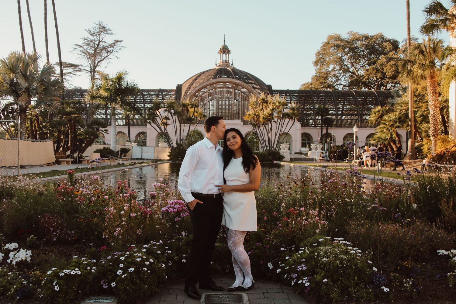 A romantic photoshoot in front of a grand botanical building with arched glass dome and blooming garden at sunset.