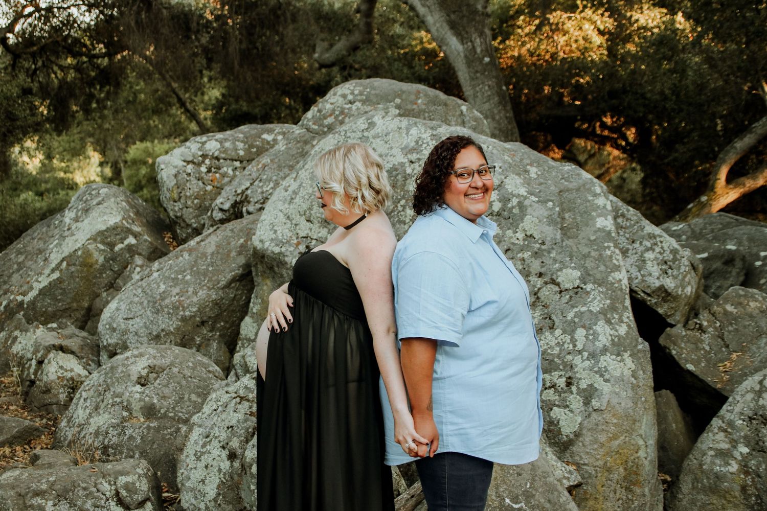 Couple poses back-to-back against large rocks in an outdoor setting during a maternity photoshoot.