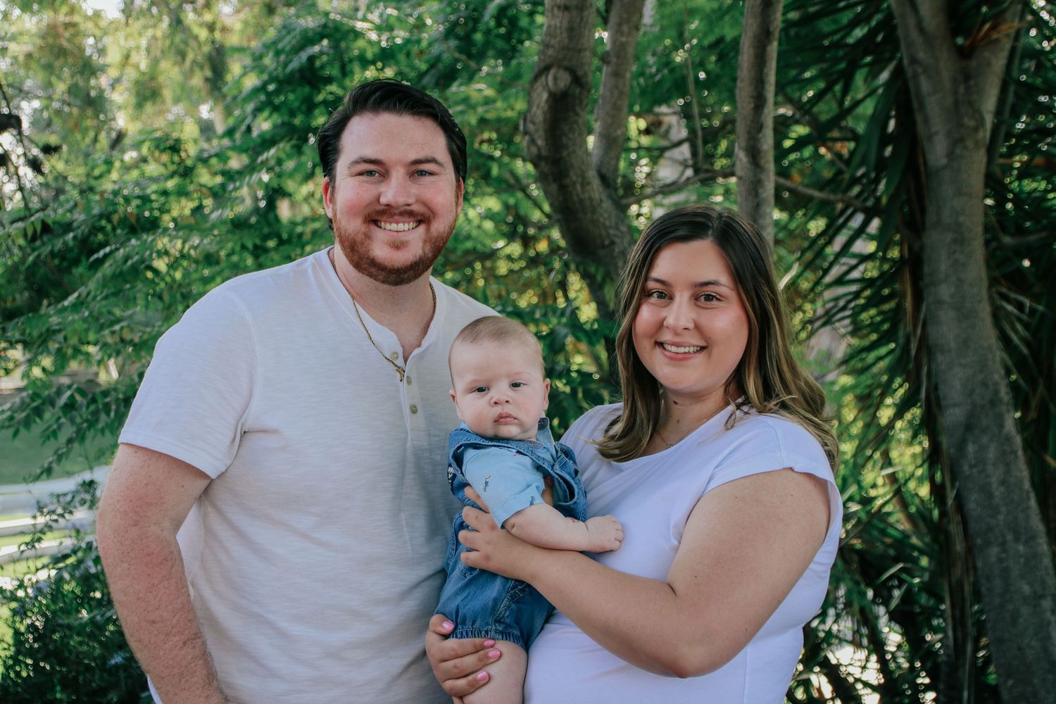 Happy family with baby poses for portrait in front of lush green trees.