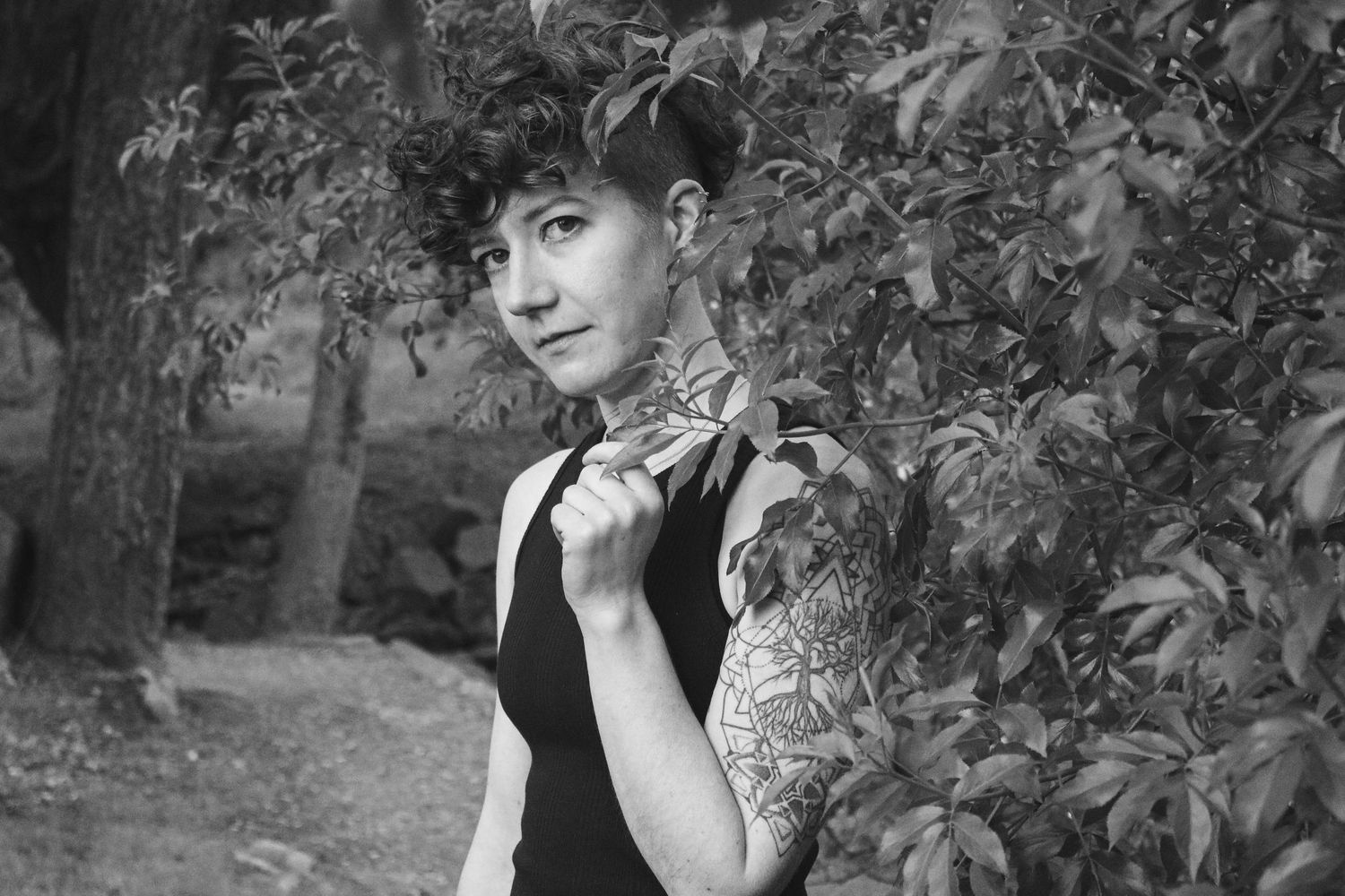 Artistic black and white portrait of a person in sleeveless black dress posed against leafy foliage outdoors.