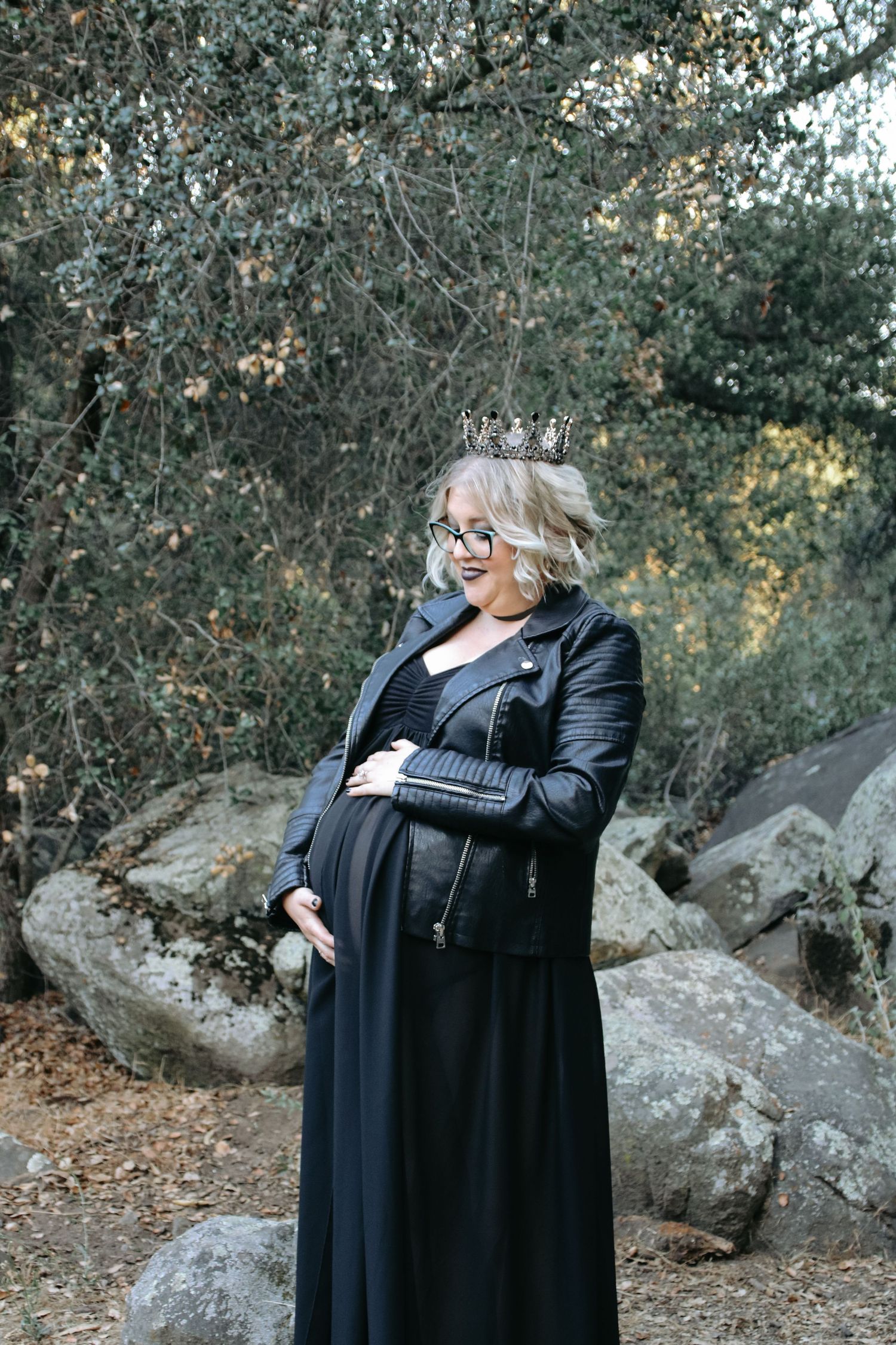 Expectant person in black dress and leather jacket poses thoughtfully among trees and rocks.