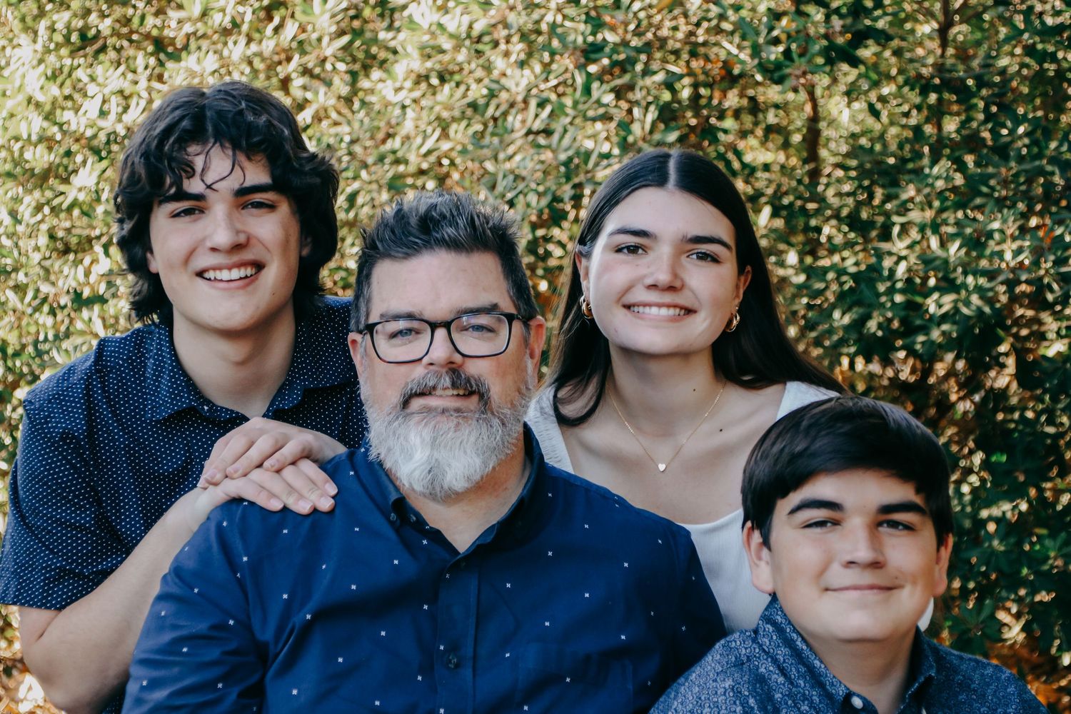 Relaxed family group photo with everyone smiling against fall foliage backdrop.