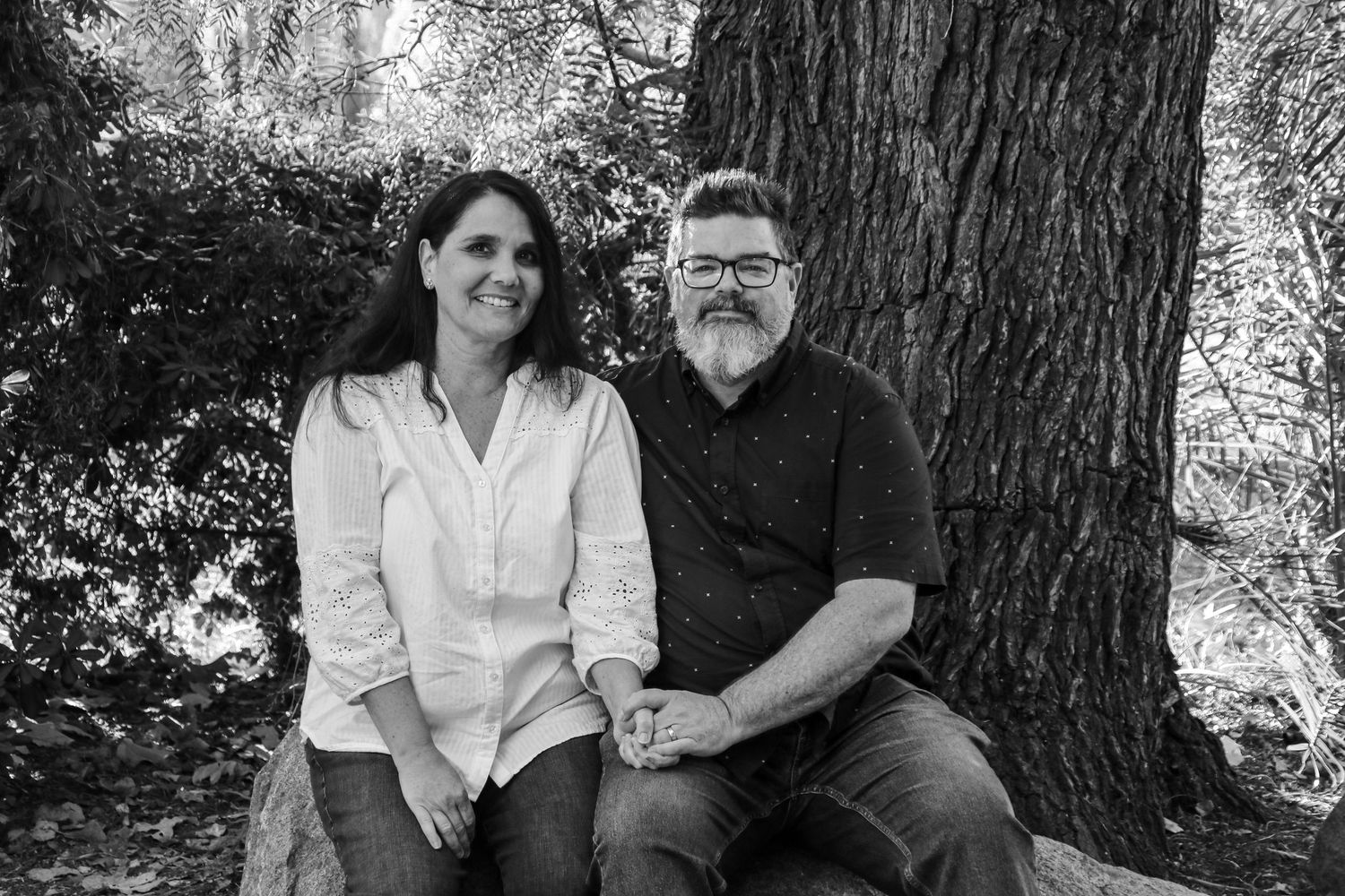 A couple sitting together on a bench under a large tree in a black and white photograph.