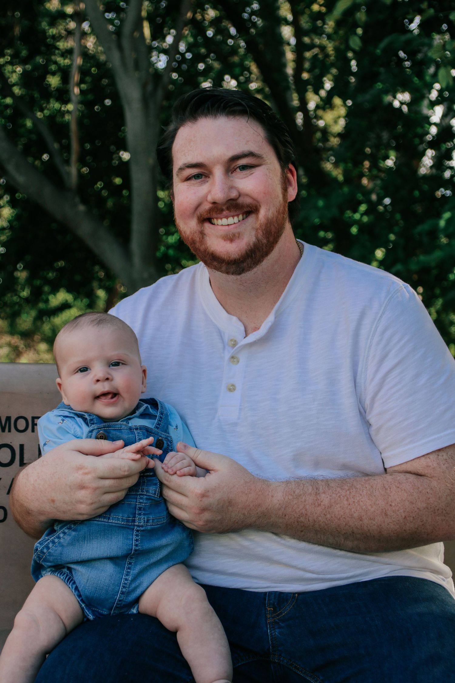 Adult and infant in denim overalls sit together outdoors on a sunny day.