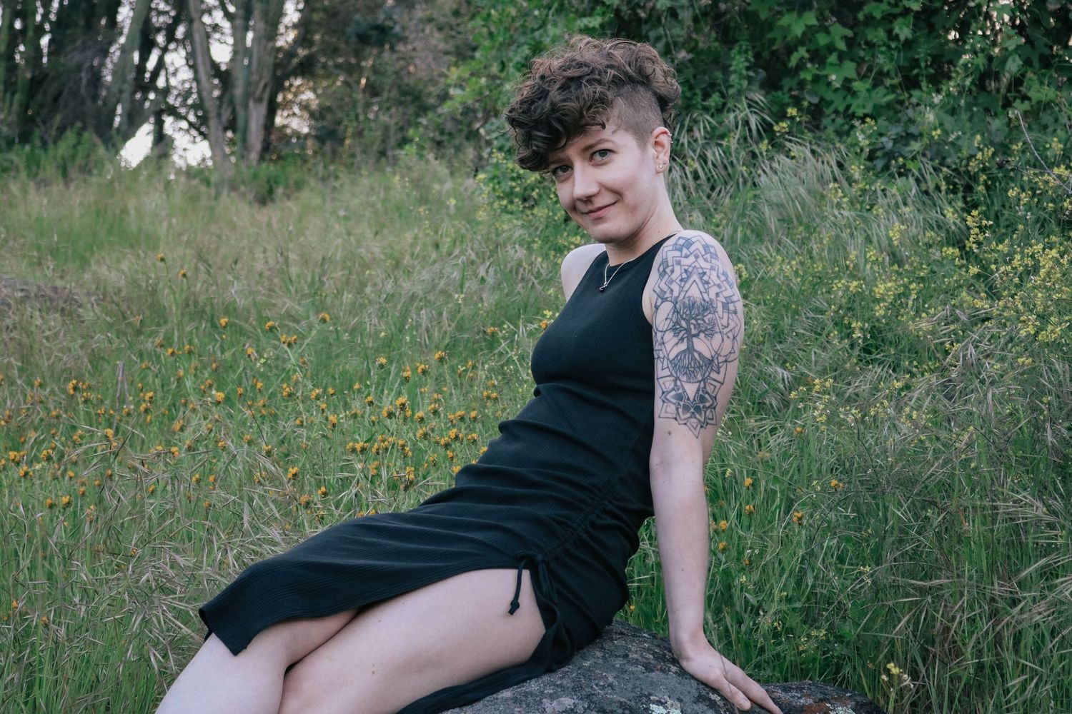 Person in sleeveless black dress poses on rock with natural backdrop.
