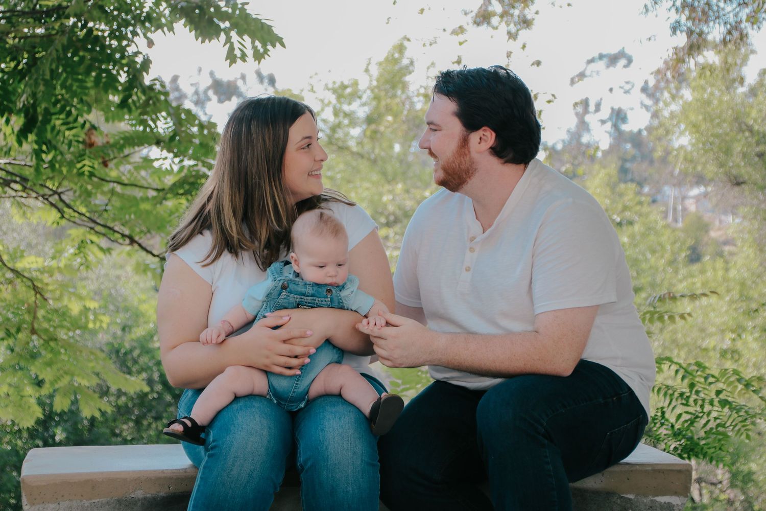 A casual outdoor family portrait with parents and baby sitting together on a wooden bench.