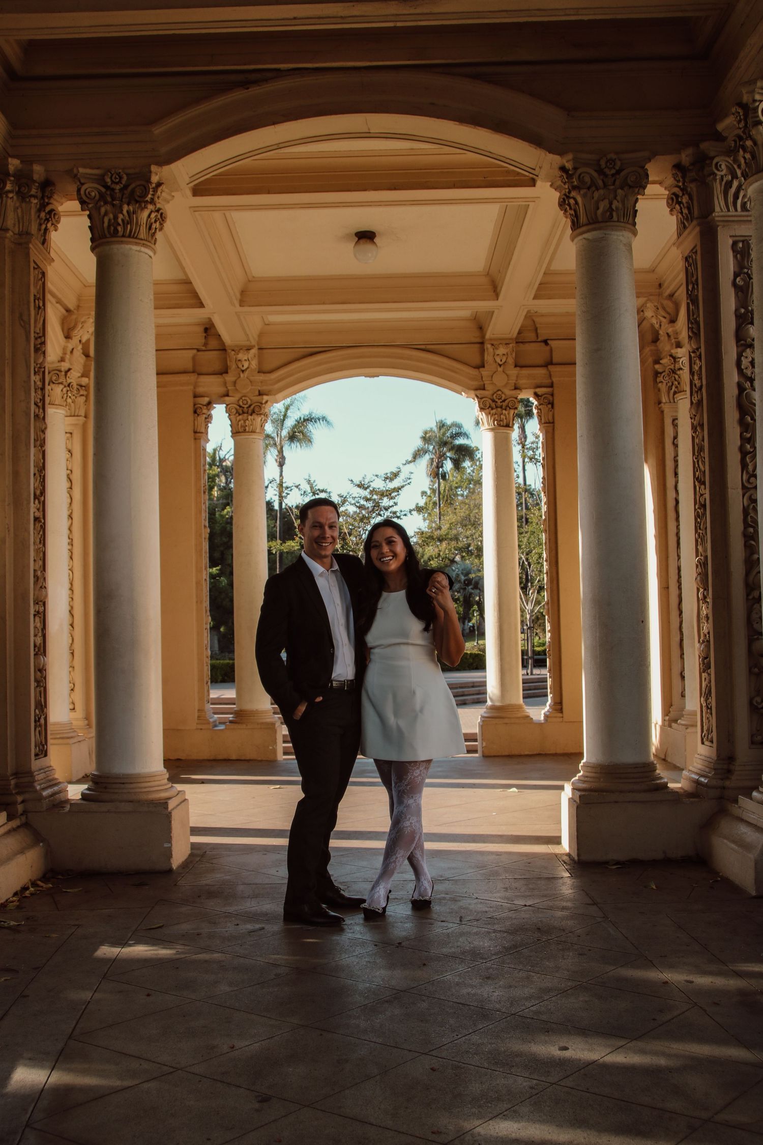 A couple poses playfully in an elegant colonnade with palm trees visible in the background.