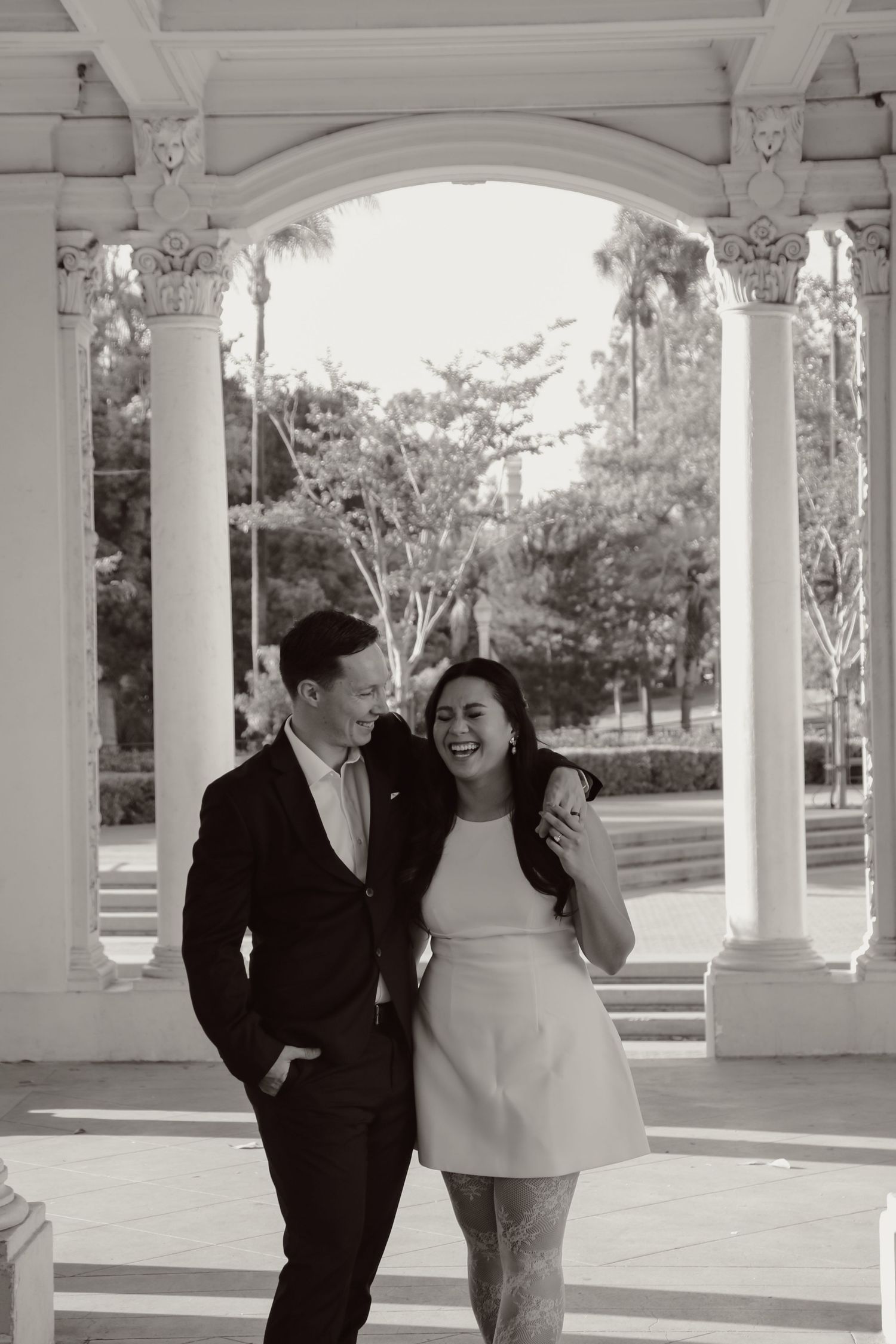 A joyful couple shares intimate moments beneath a white columned pavilion in a park setting.