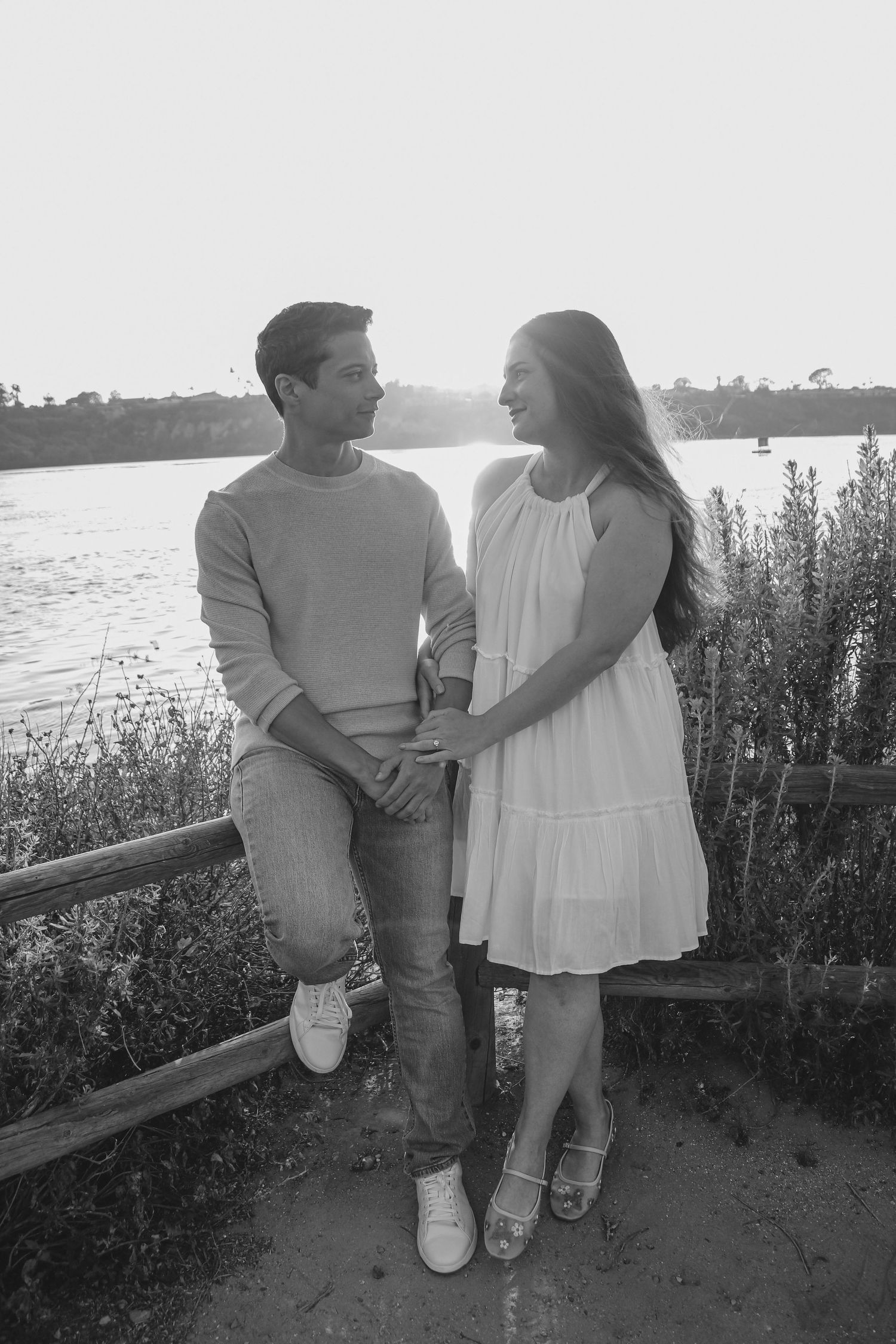 Black and white photo of couple sitting on railing by waterfront in casual attire at sunset.