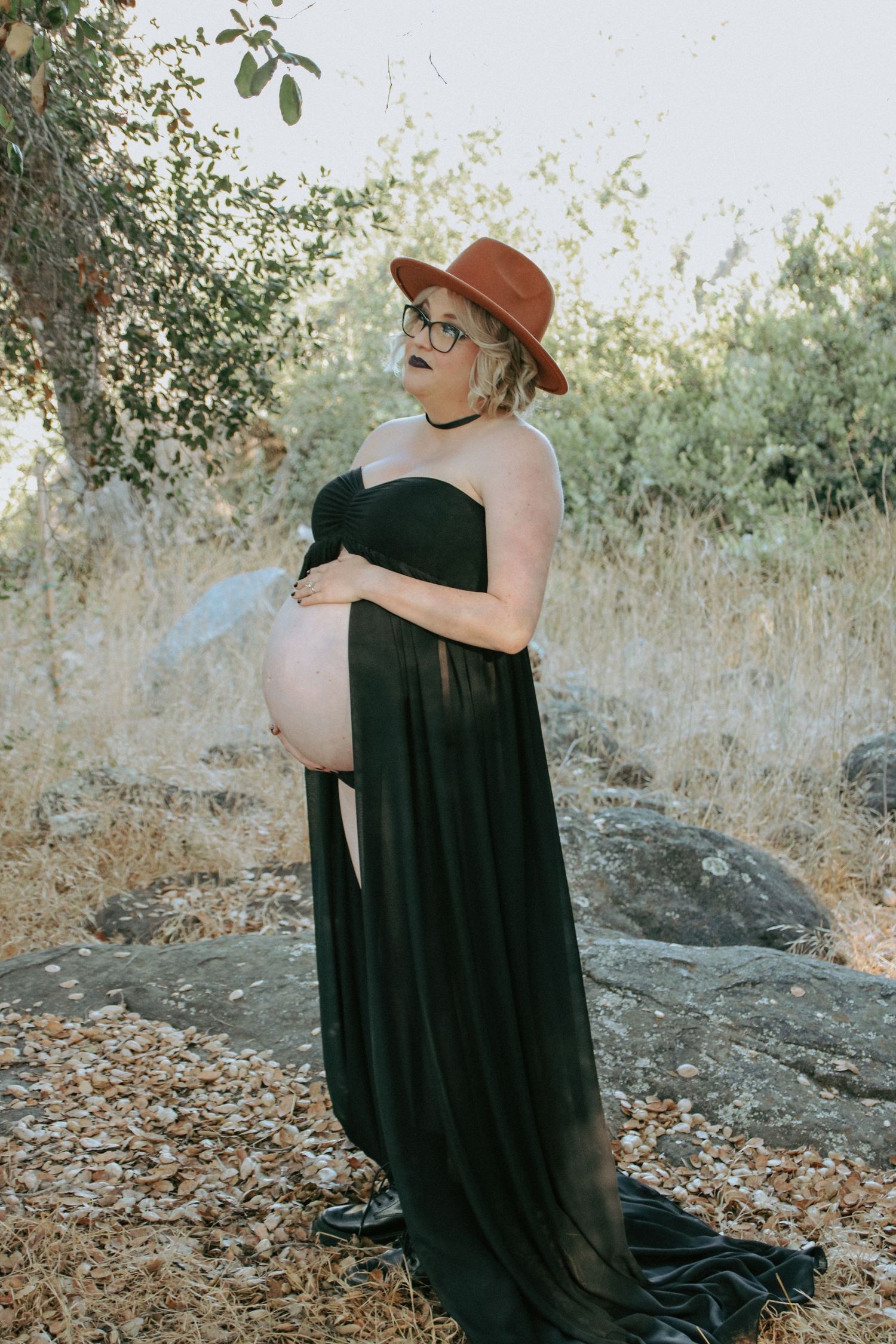A maternity photoshoot in nature features an elegant black dress and brown hat against a sunlit outdoor background.