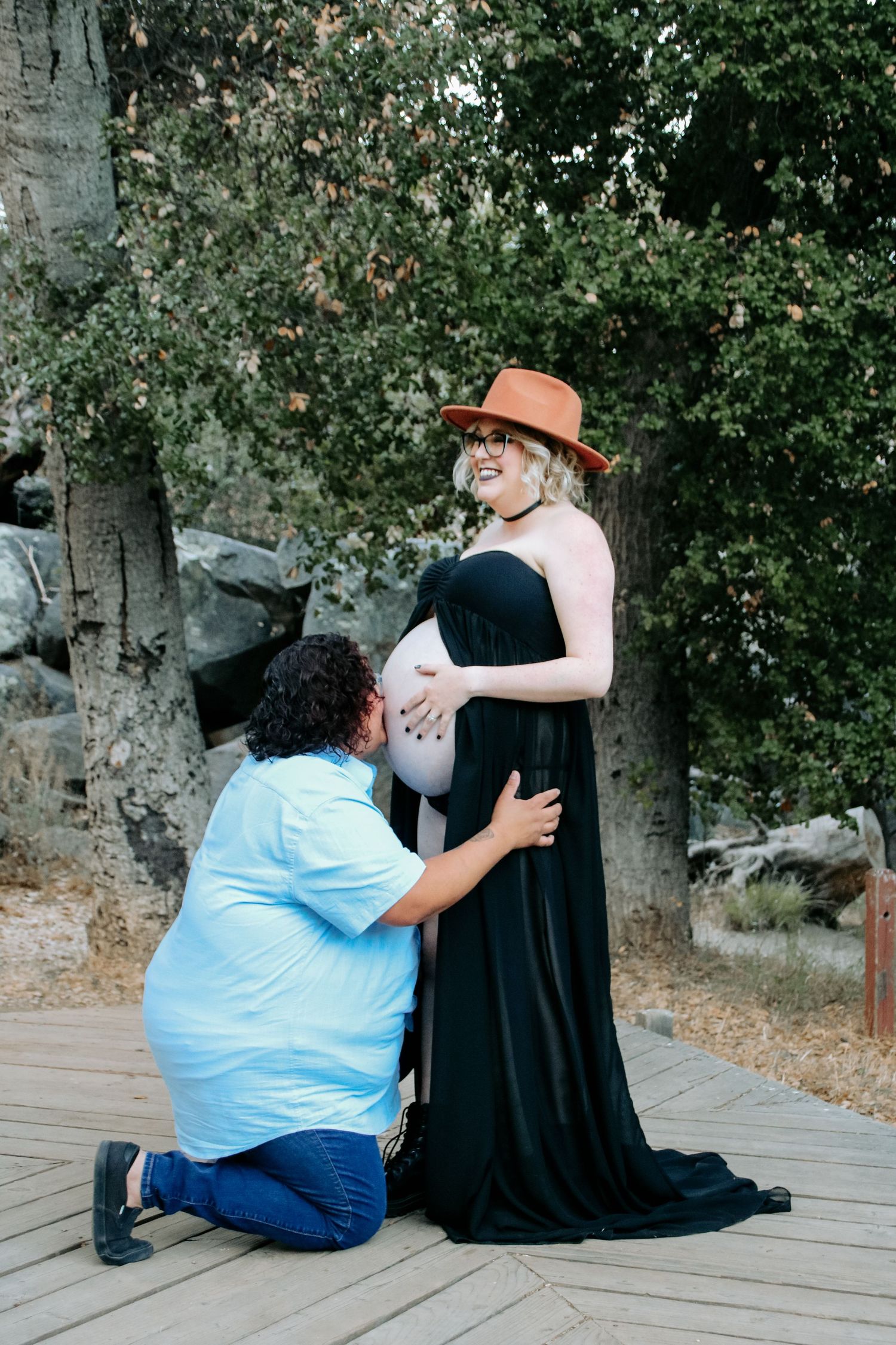 A maternity photo shoot outdoors shows a partner kneeling and touching a pregnant person's belly who wears a black dress and orange hat.