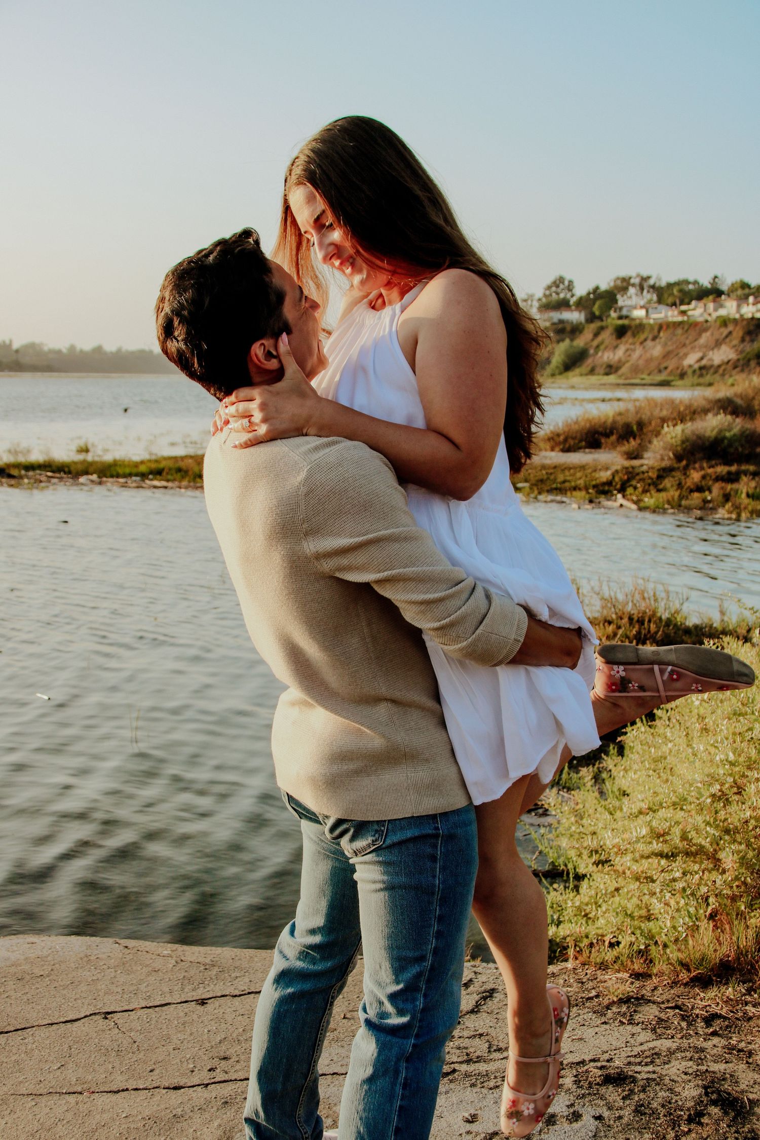 A couple embracing in a white dress and beige sweater against a scenic waterfront backdrop.
