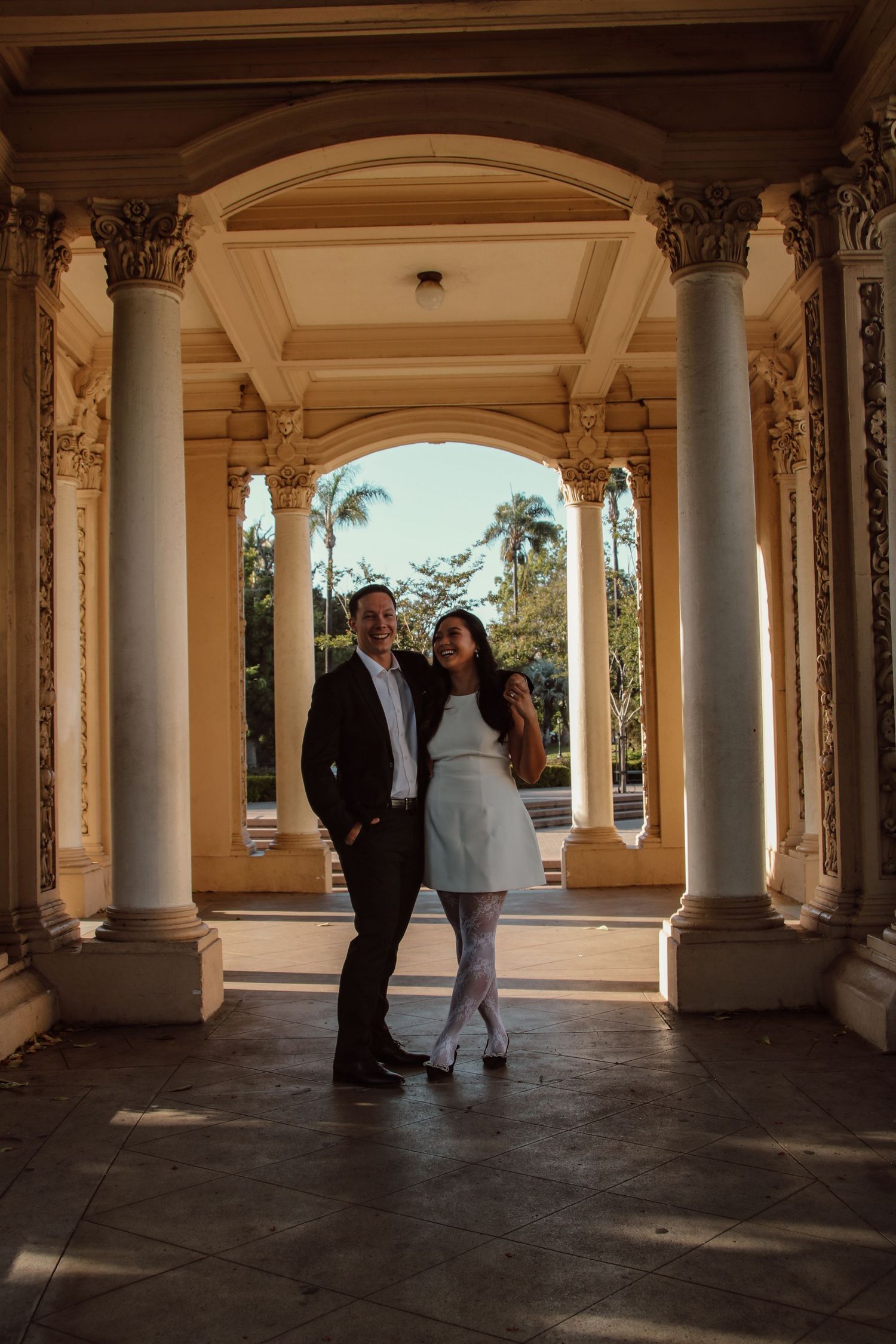 A couple interacts beneath classical architecture with sunlit palm trees framed by arches.