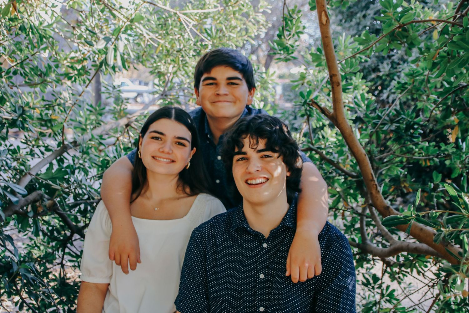 Three people posing together outdoors under green foliage in casual attire.