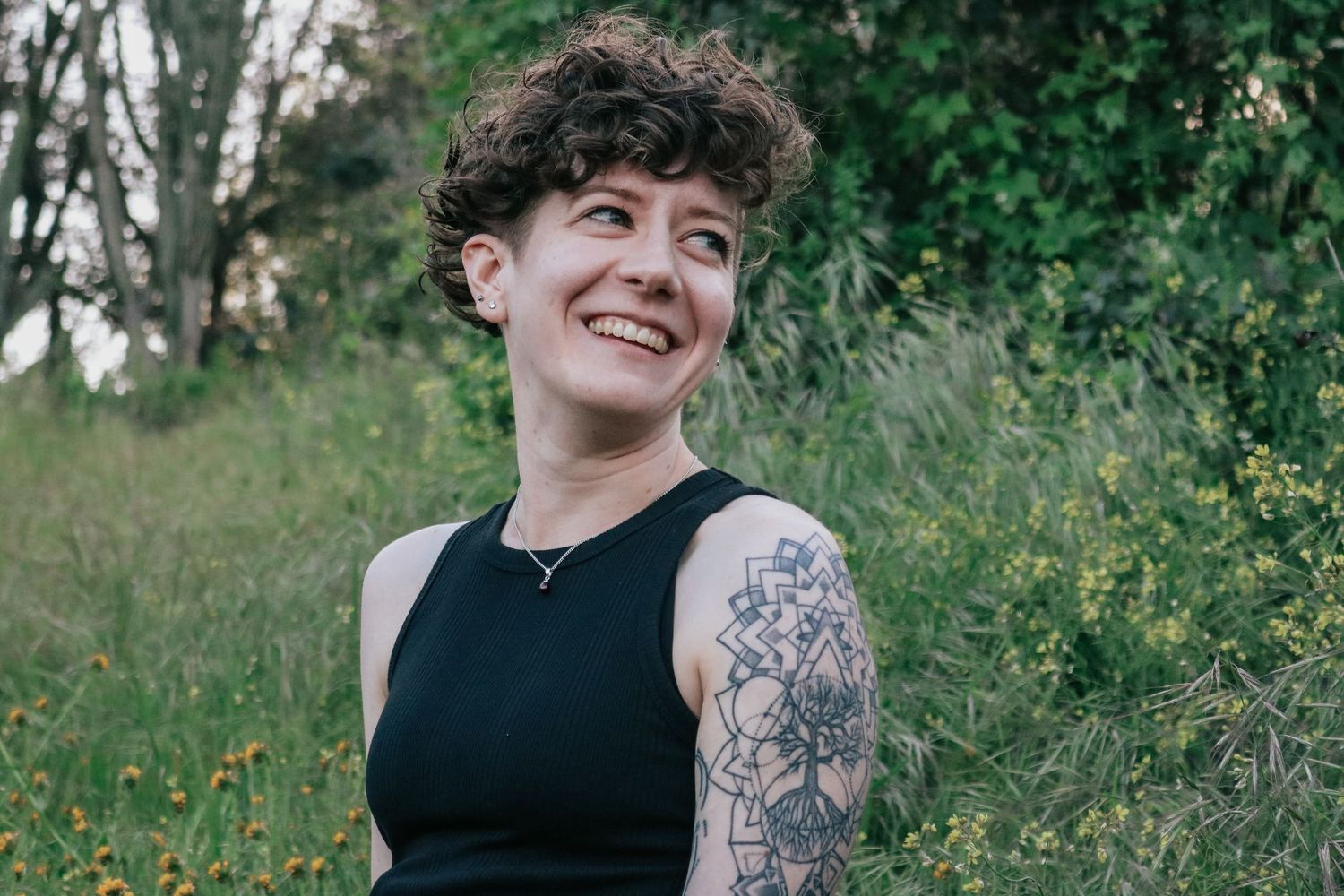 Person with curly hair and tattoo sleeve smiles while sitting on rock outdoors.