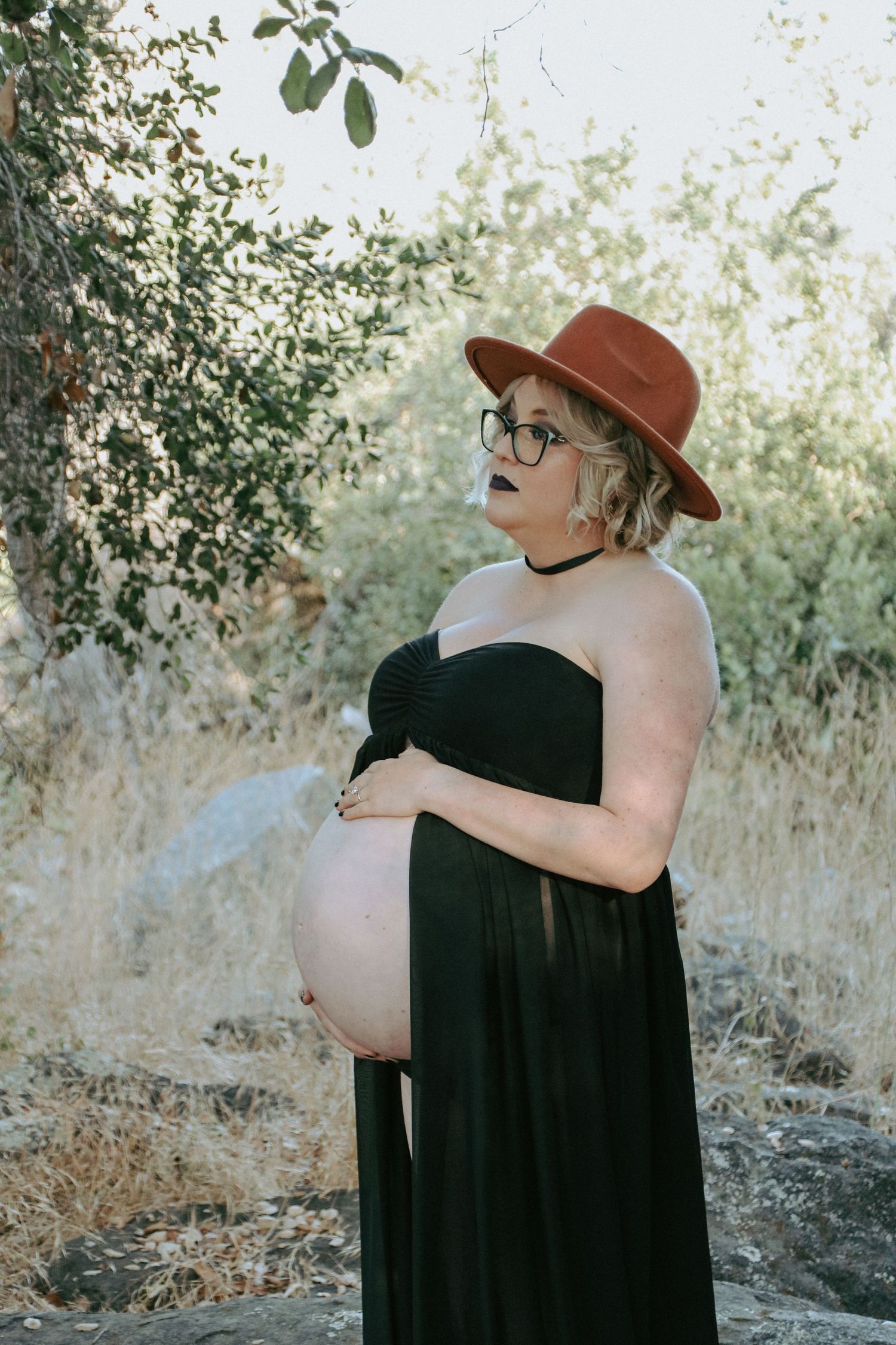 A maternity photo in nature showing an expectant mother in a black dress and brown hat against a lush outdoor backdrop.
