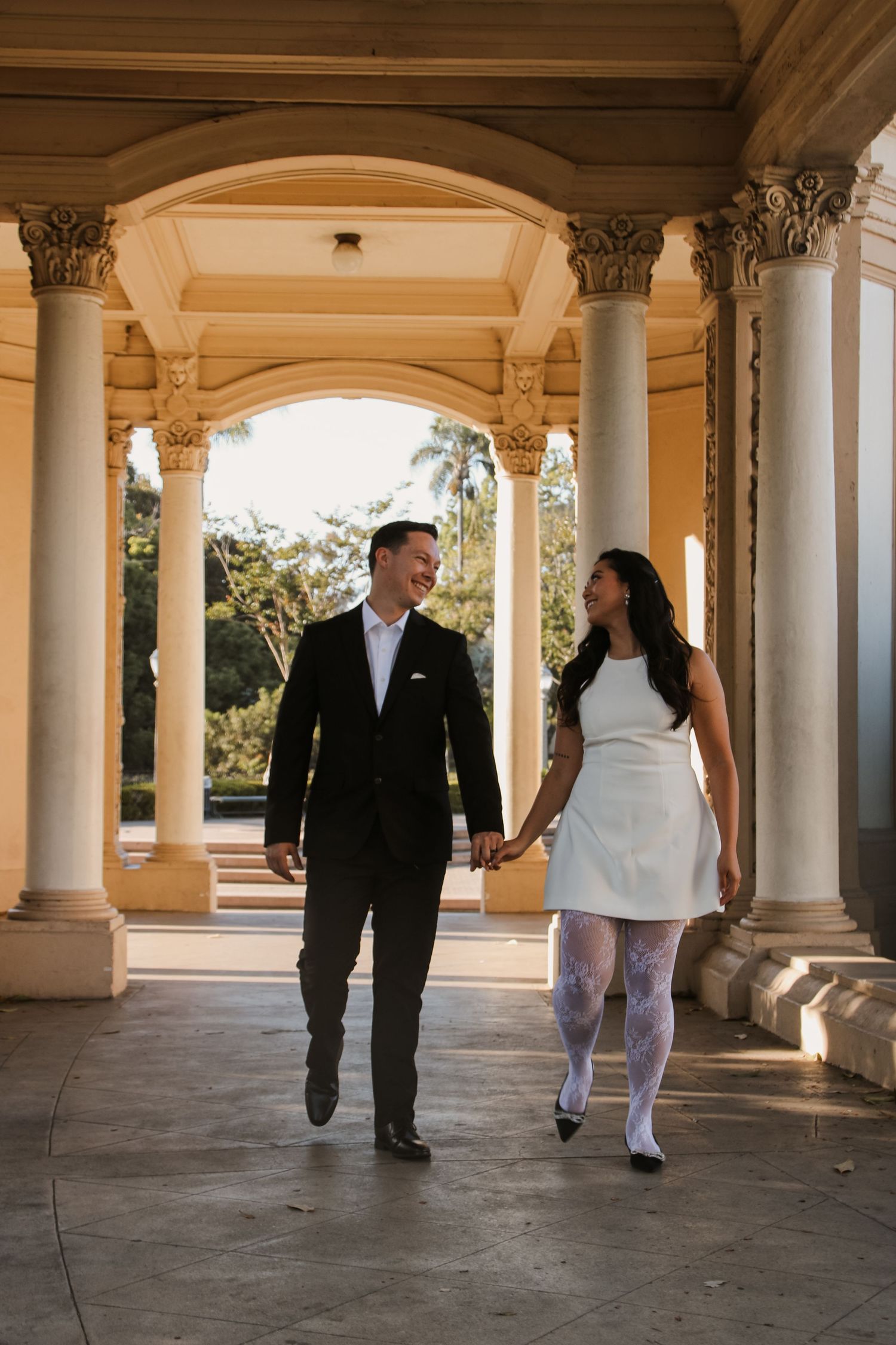 Couple in formal attire walking through sunlit colonnade with decorative pillars.