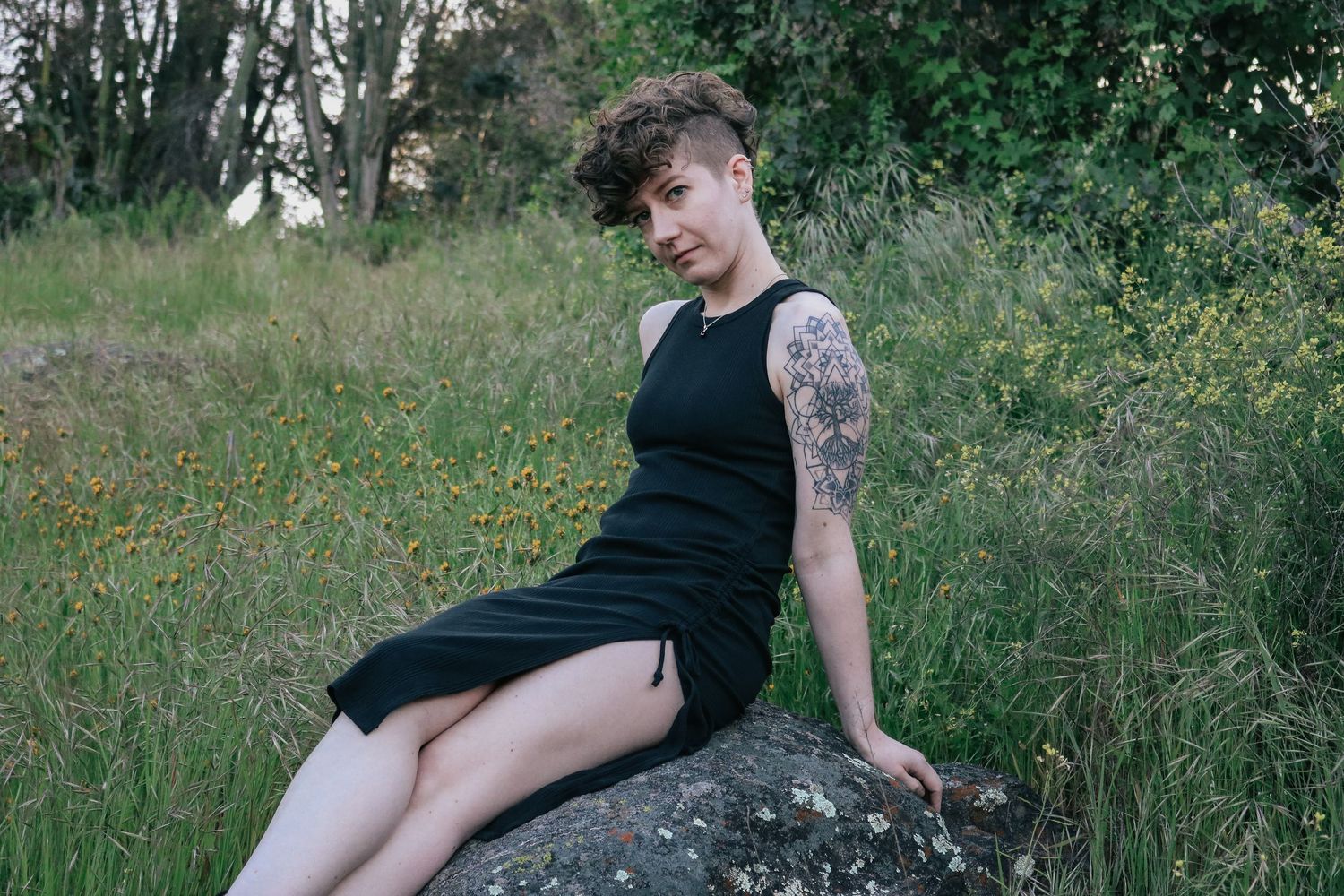 Person in black dress sits casually on rock in outdoor garden setting.
